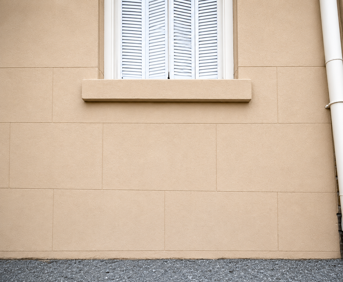 Beige stucco wall with a small window and white shutters above a gravel strip