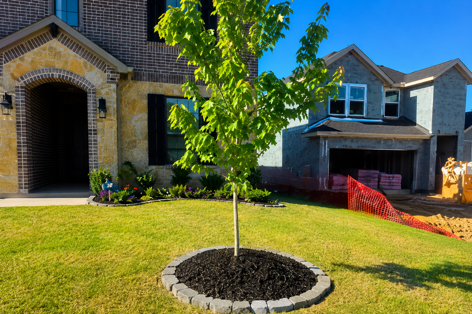 Front yard with a young tree, green lawn, and a brick house with a garage in the background