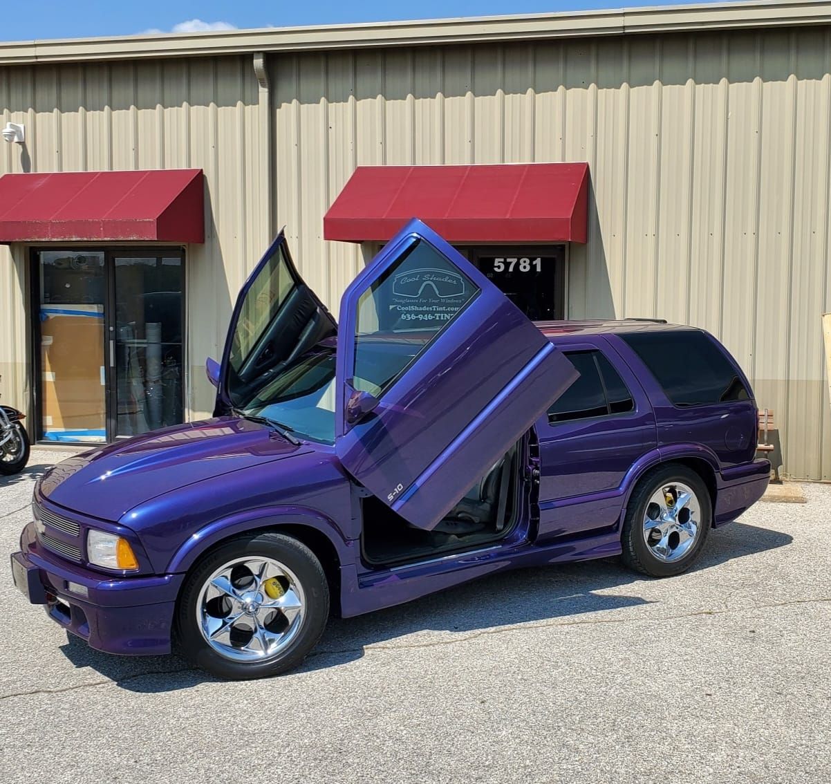 Purple sports car with scissor doors open, parked outside a beige building with red awnings.