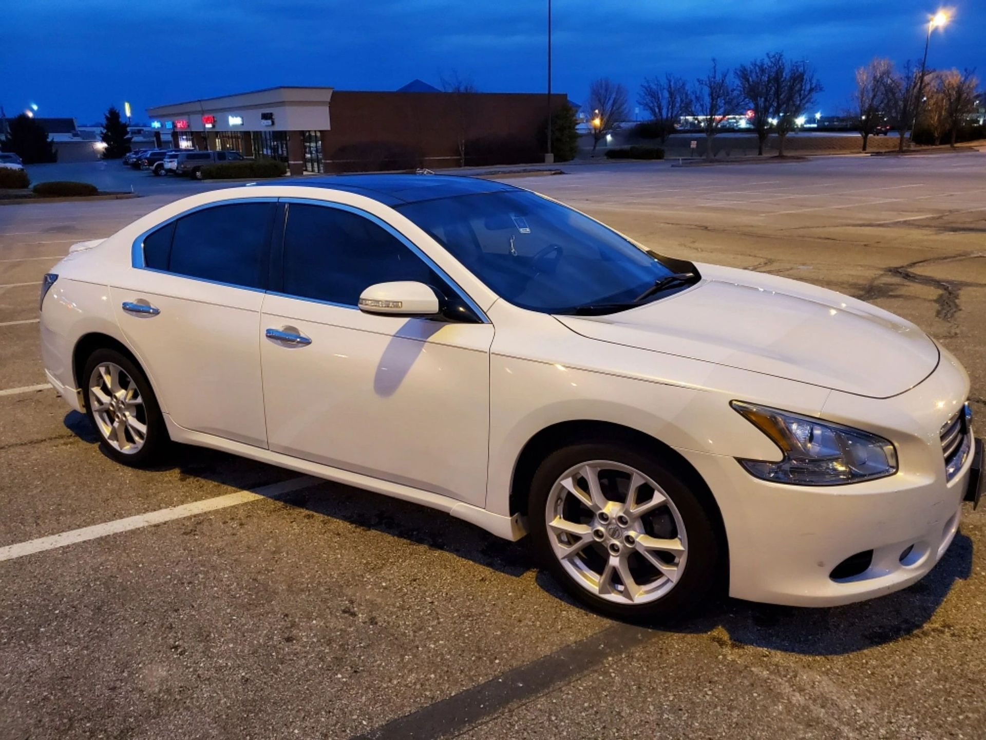 White sedan parked in a lot at dusk with storefronts in the background
