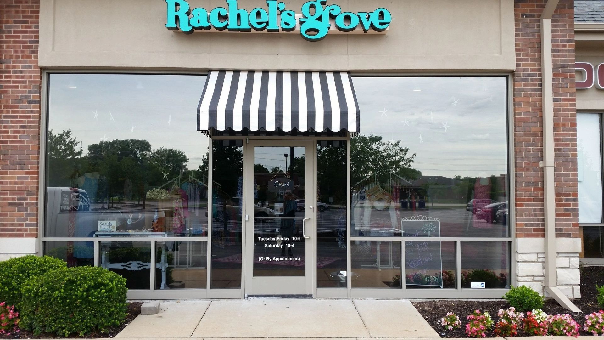Storefront with black-and-white striped awning and glass doors, labeled “Rachel’s Gown,” with flowers out front
