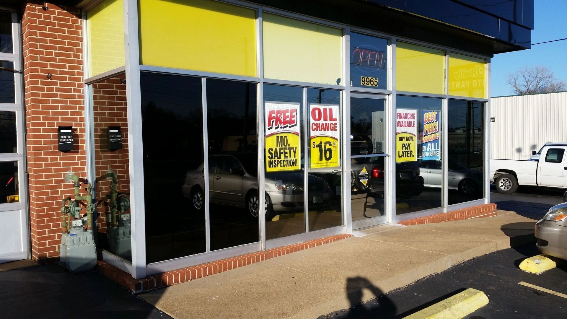 Storefront with large dark windows and yellow sale signs, viewed from the sidewalk on a sunny day