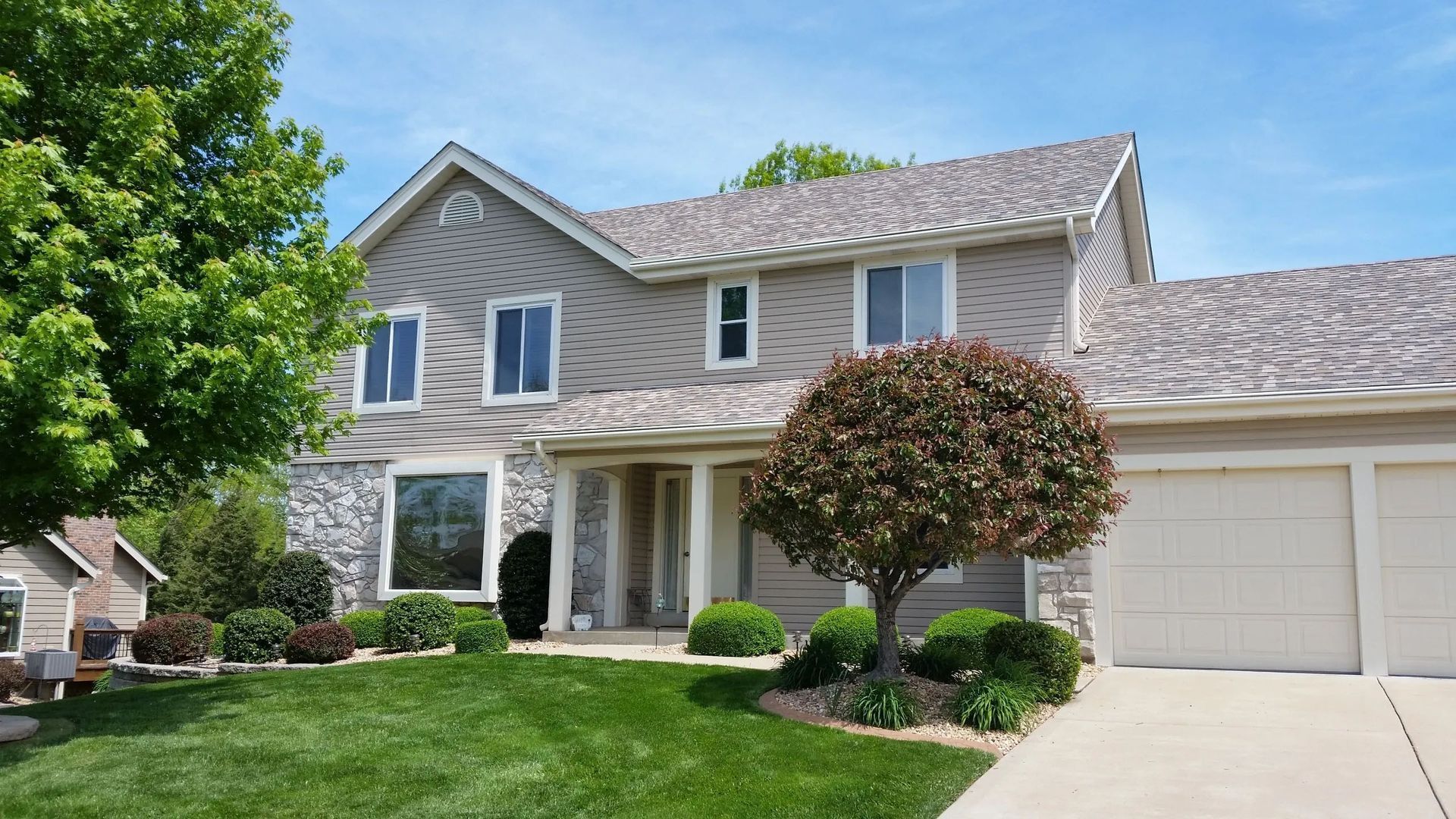 Suburban two-story house with beige siding, white garage, front porch, and manicured green lawn under blue sky