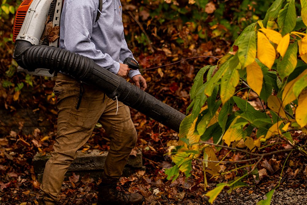 Man using a leaf blower, brown pants, blue shirt, fall leaves.