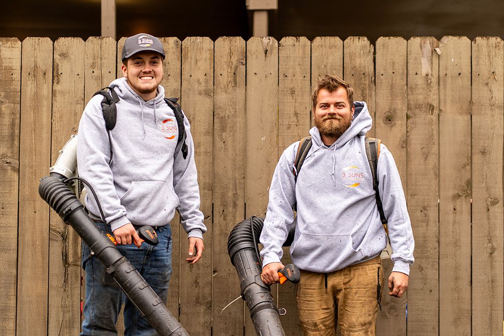 Two men wearing gray hoodies and backpacks, holding leaf blowers in front of a wooden fence.