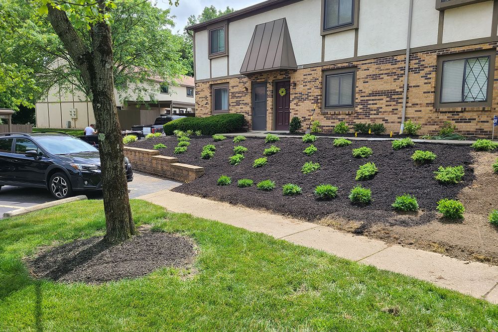 A two-story building with freshly mulched garden beds, green plants, and a black SUV parked nearby.