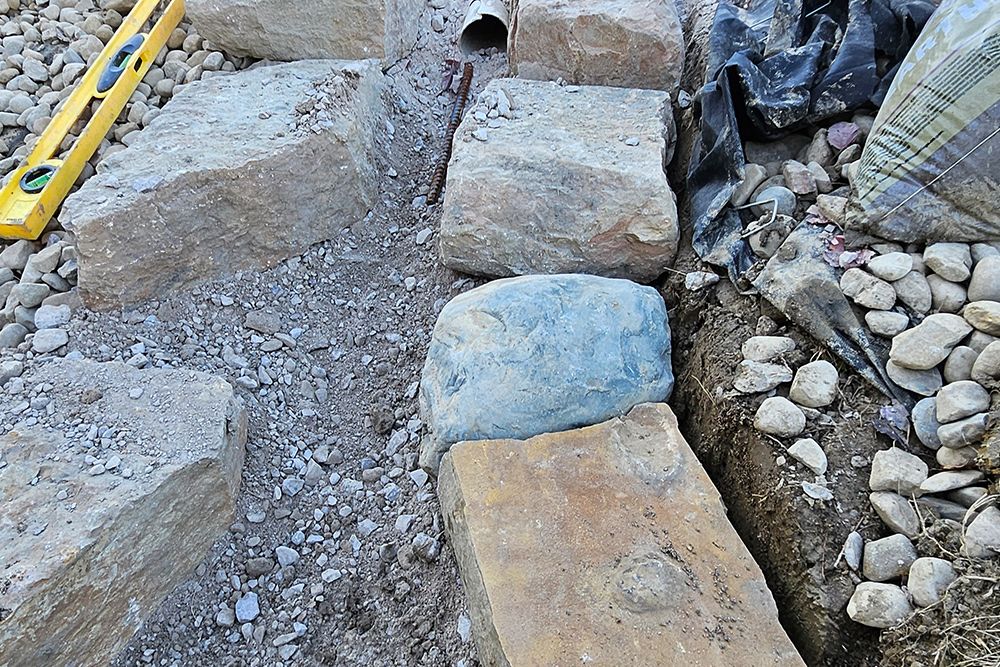 Stones arranged for construction. A level and pipe are visible. Gravel and dirt are present.