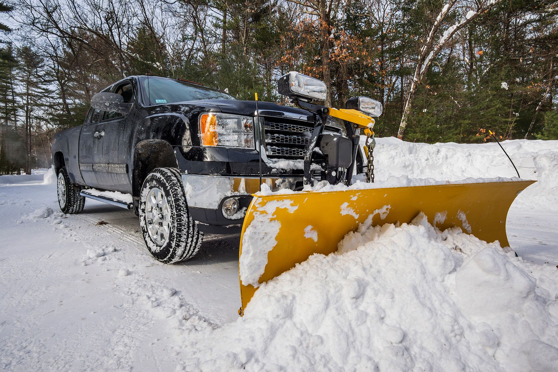 Black truck with yellow snowplow clearing a snow-covered driveway in a wooded area.