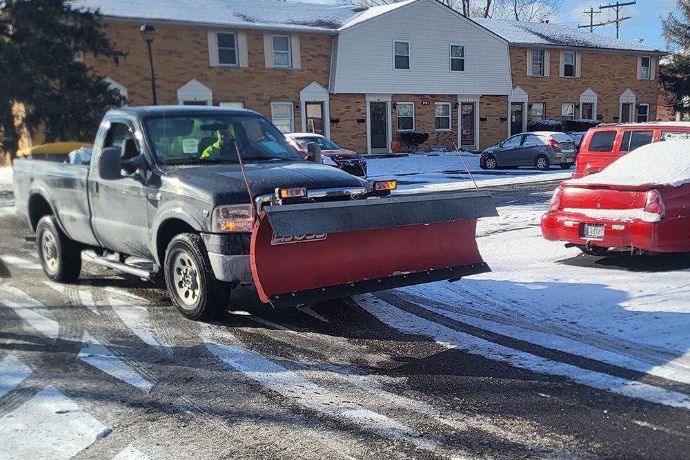 Gray pickup truck with snowplow clearing snow from a street. Snow-covered townhouses in the background.