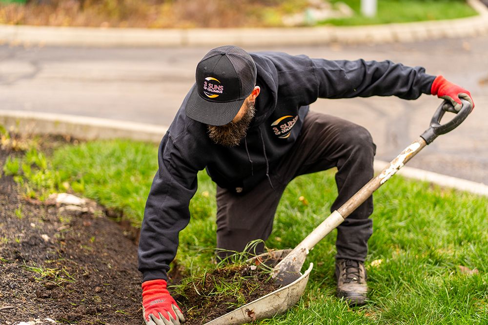 Man kneeling, digging in dirt with a shovel, wearing black hoodie, cap, and red gloves outdoors.
