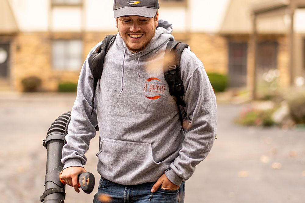 Man using a leaf blower, wearing a gray hoodie and hat, smiling outside a building.