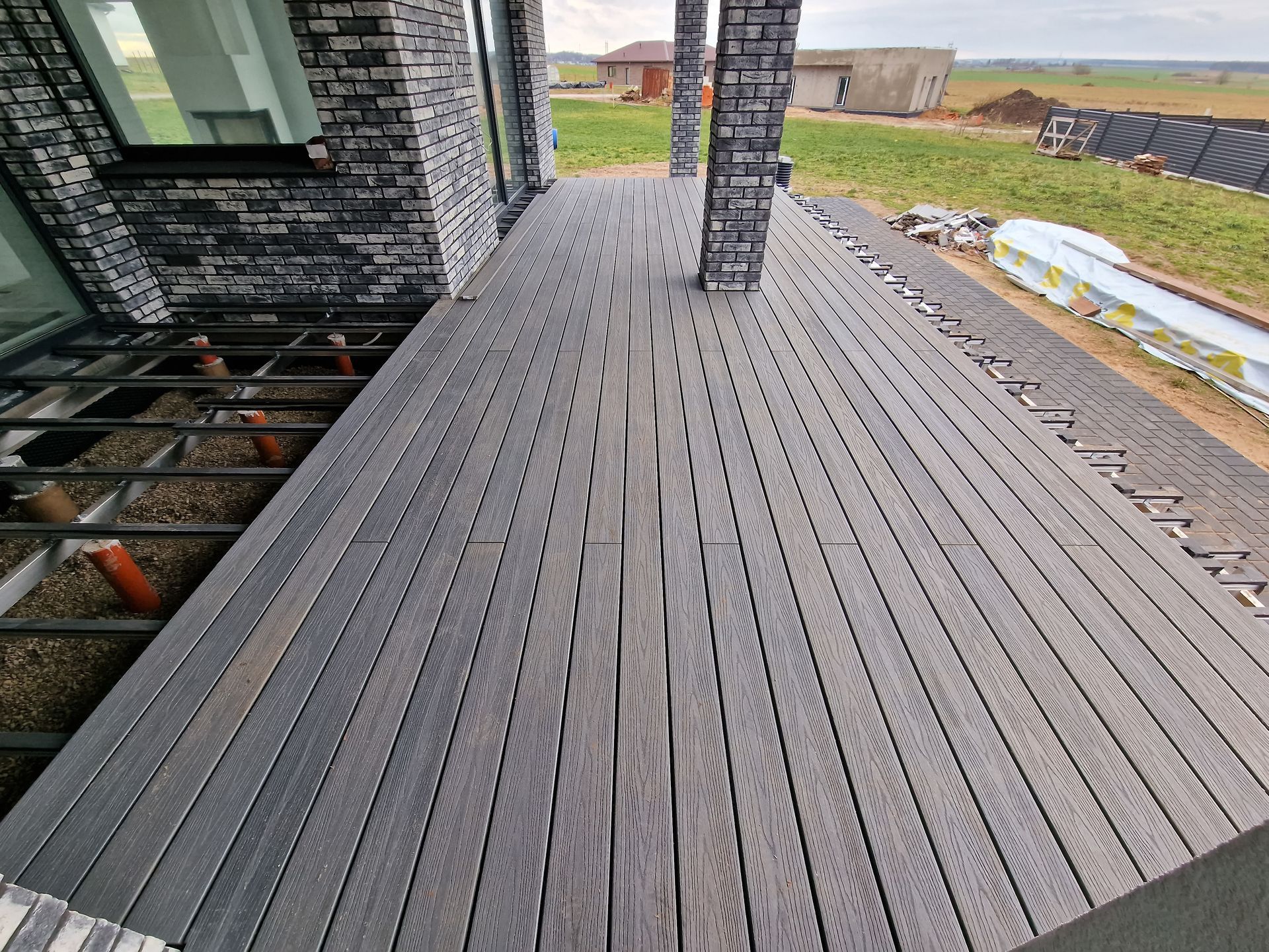 Une terrasse extérieure partiellement aménagée, avec des planches de bois foncé installées à côté d'une structure en acier apparente adossée à une maison en briques.