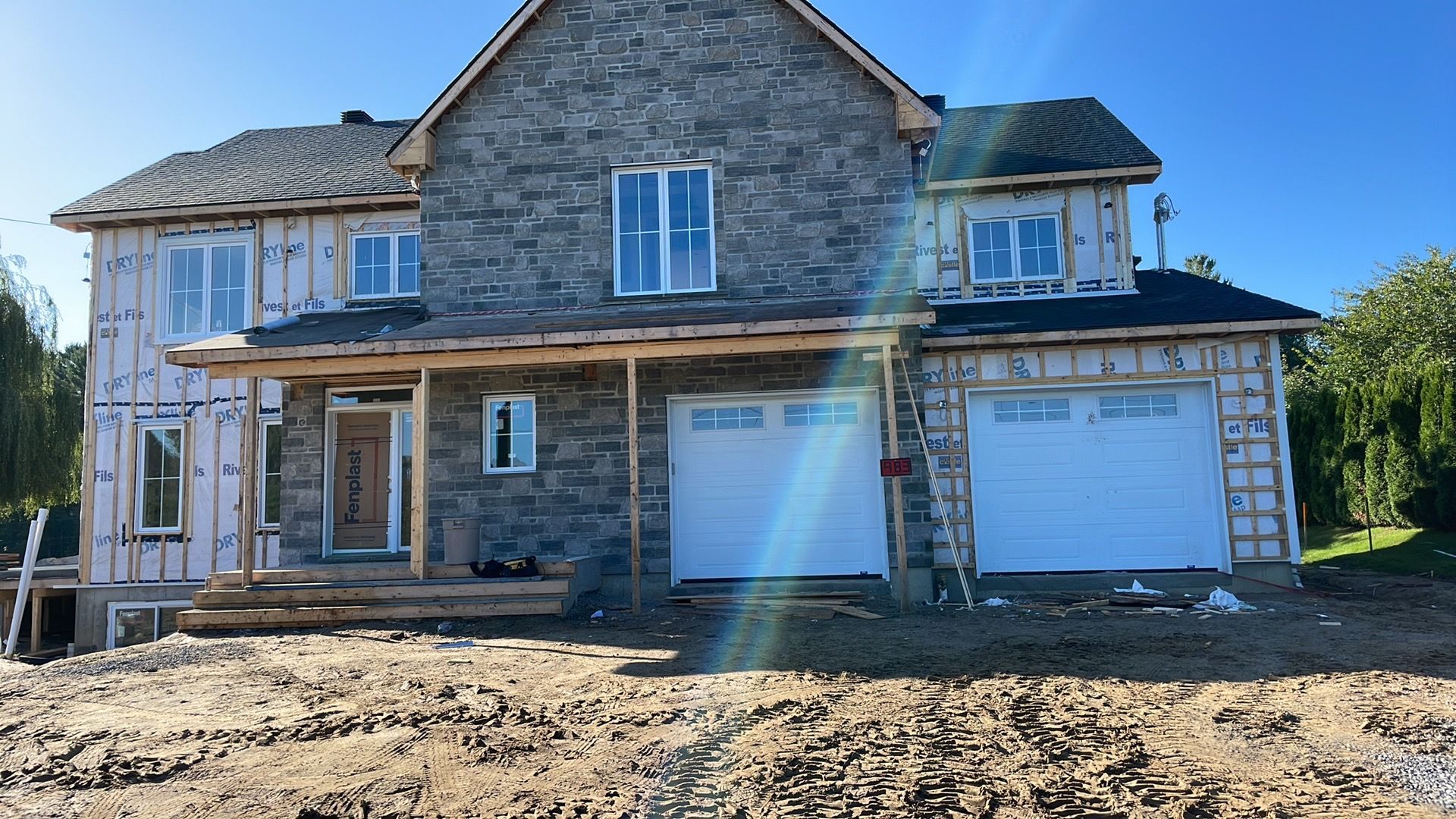 Une maison à deux étages en construction, avec une façade en pierre au centre, des panneaux isolants roses et deux portes de garage blanches.