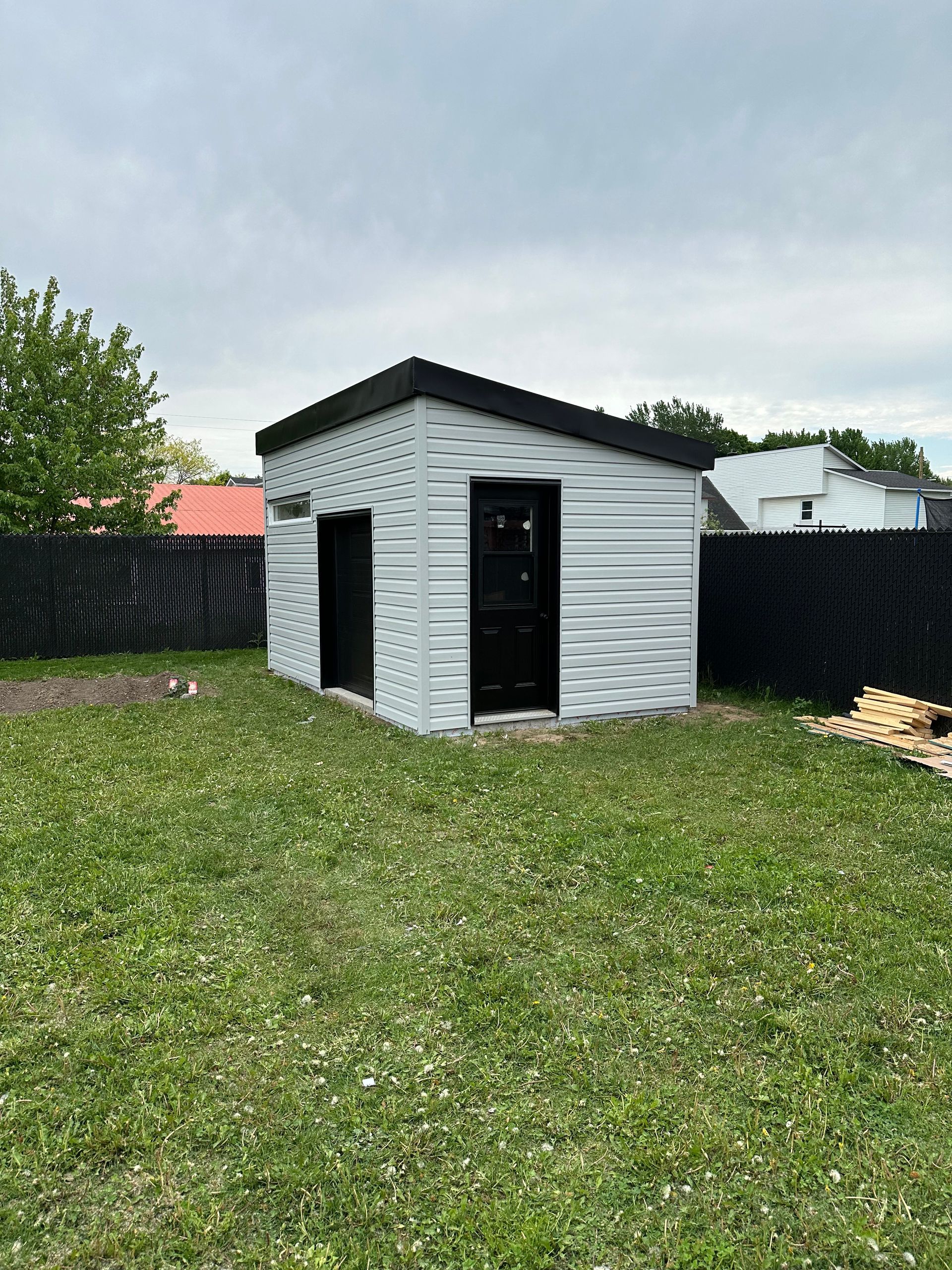 Un cabanon blanc avec une porte et un toit noirs se dresse dans une cour herbeuse, entourée d'une clôture noire pour préserver l'intimité, sous un ciel nuageux.