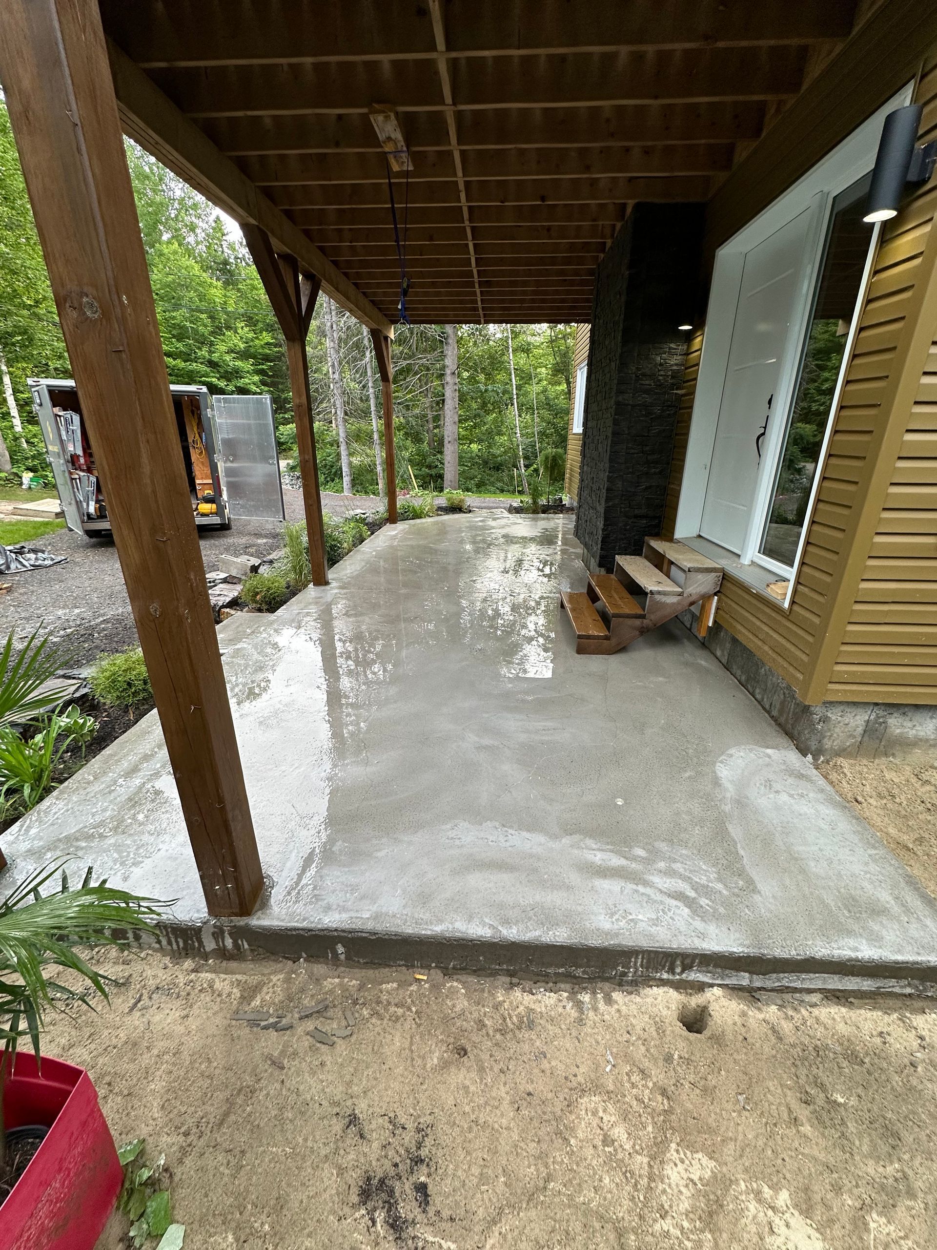 Une terrasse en béton fraîchement coulée sous un toit de porche en bois, adjacente au mur extérieur d'une maison avec des marches.