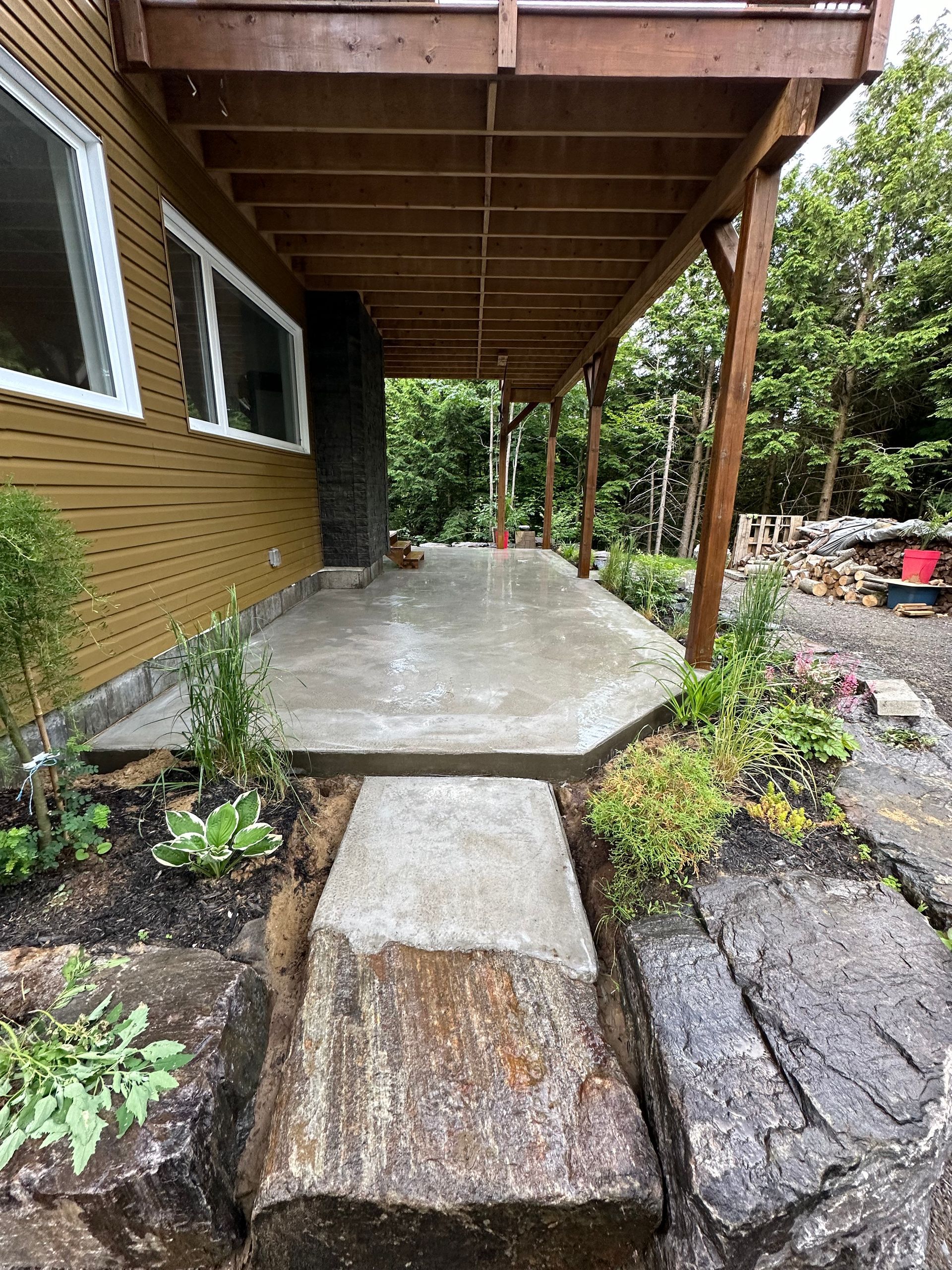Une terrasse en béton fraîchement coulée sous une terrasse en bois, reliée par une marche en pierre, adossée à une maison au bardage jaune.