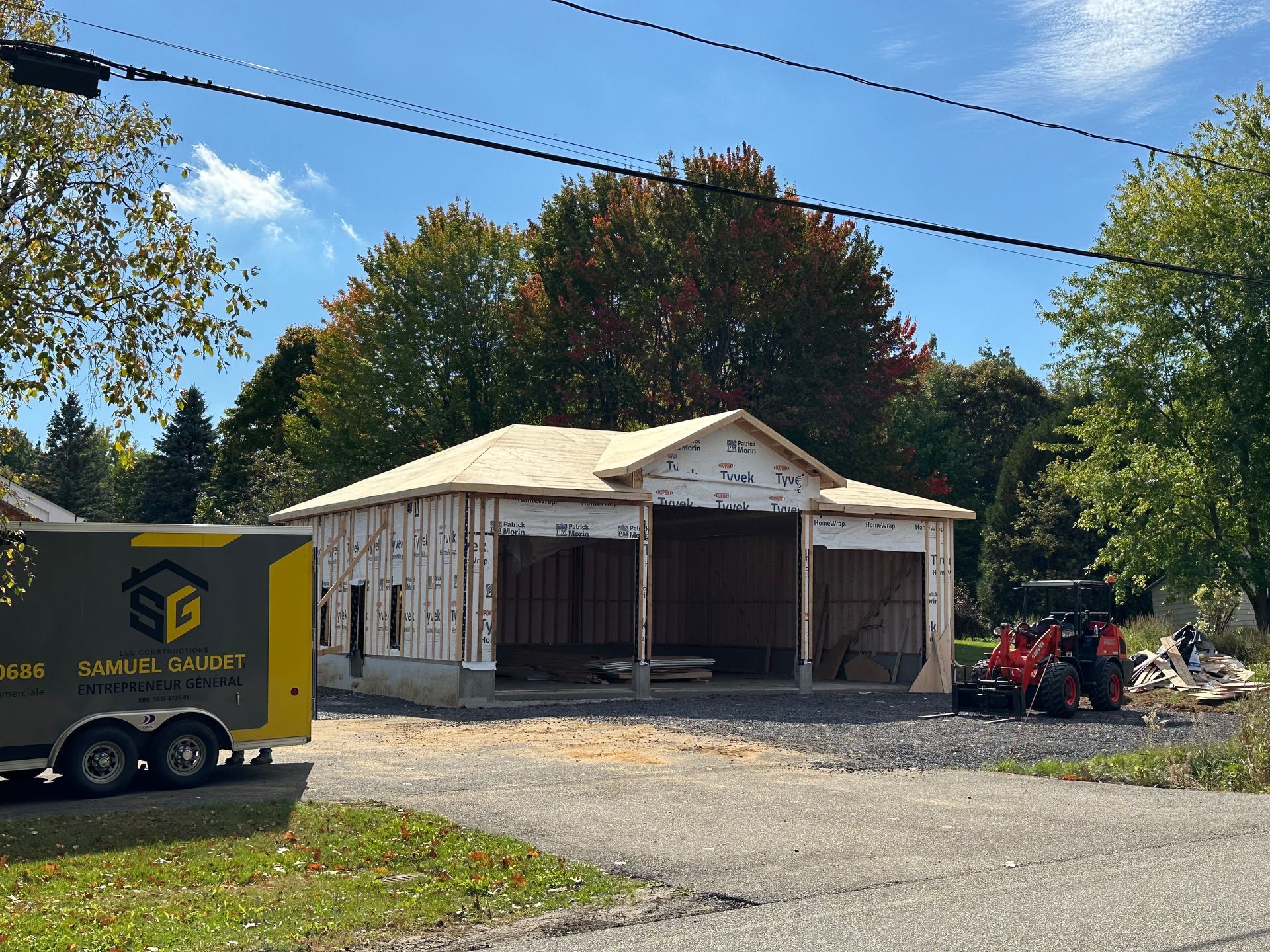 La structure d'une maison neuve est en construction, avec une remorque d'entreprise à gauche et un tracteur rouge à droite.