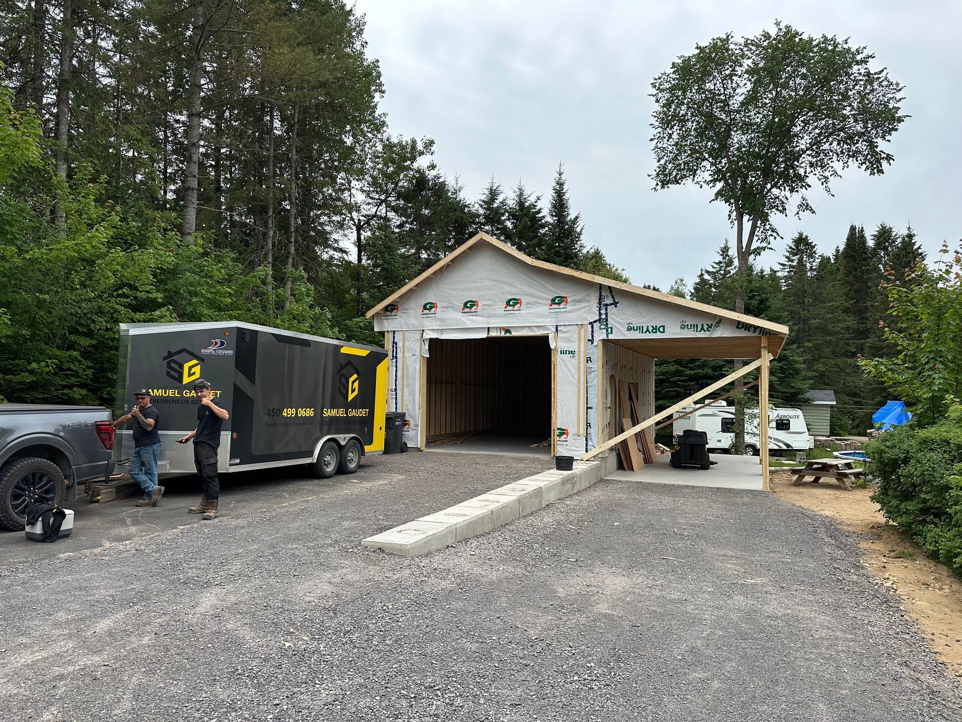 Deux personnes se tiennent près d'une camionnette grise et d'une remorque noire, à côté d'un garage en construction.
