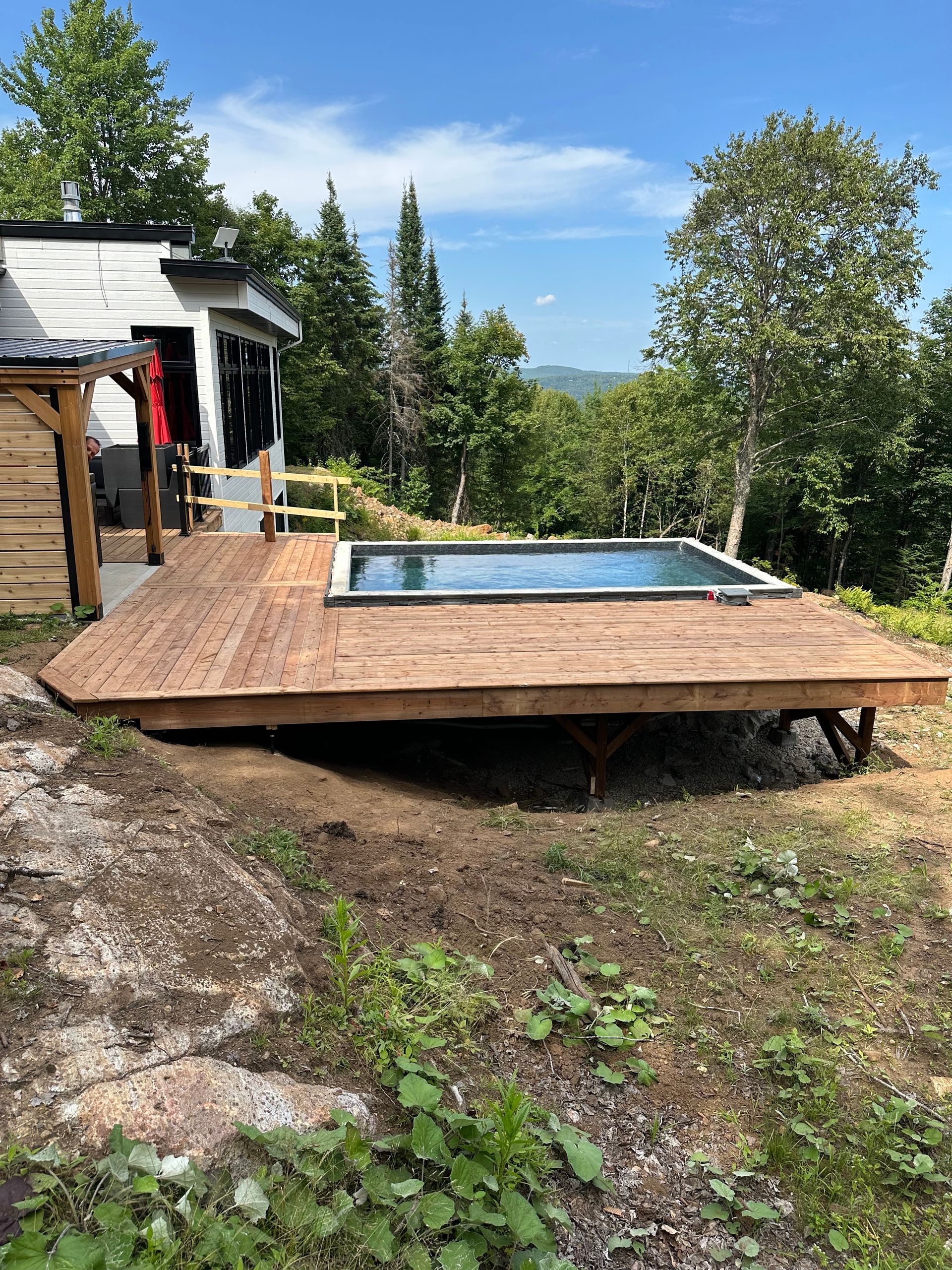 Une piscine hors sol rectangulaire aménagée sur une terrasse en bois, jouxtant un chalet blanc et donnant sur une forêt par une journée ensoleillée.