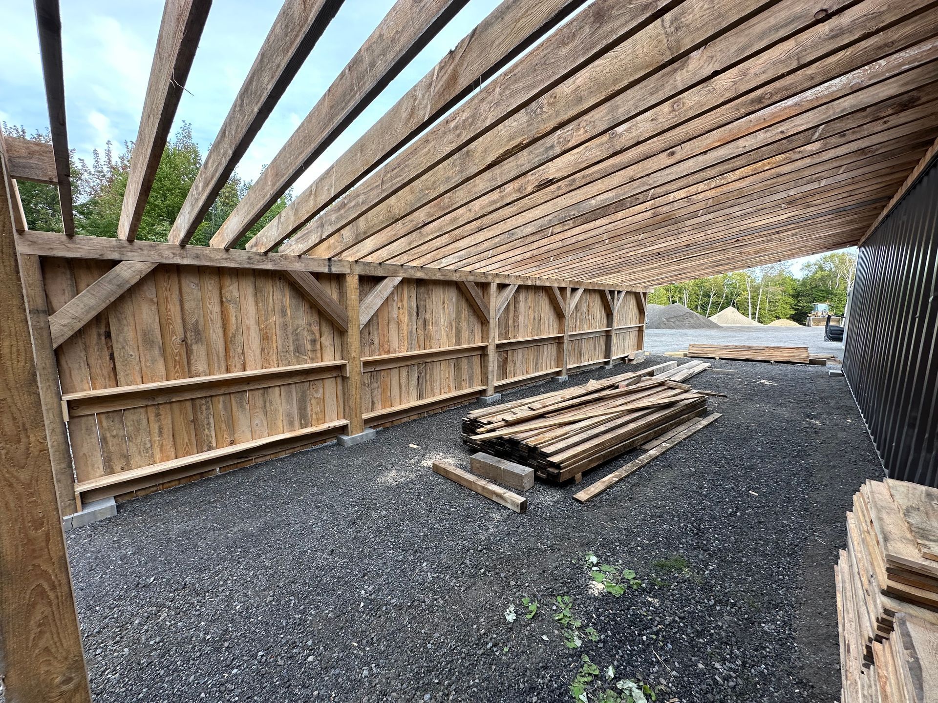 Une structure extérieure à ossature bois avec un toit en bois en pente et un sol en gravier, adjacente à un mur en tôle ondulée sombre.