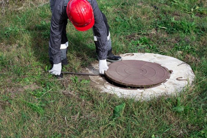 Worker in red hard hat lifting a manhole cover in a grassy area.