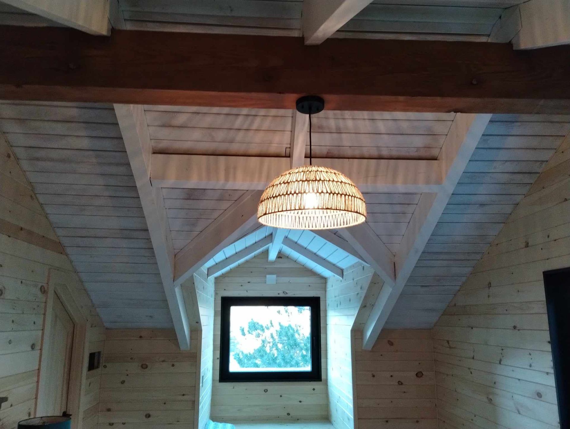 Pièce mansardée lambrissée avec plafond voûté, suspension lumineuse en tissu tressé et petite fenêtre au fond.