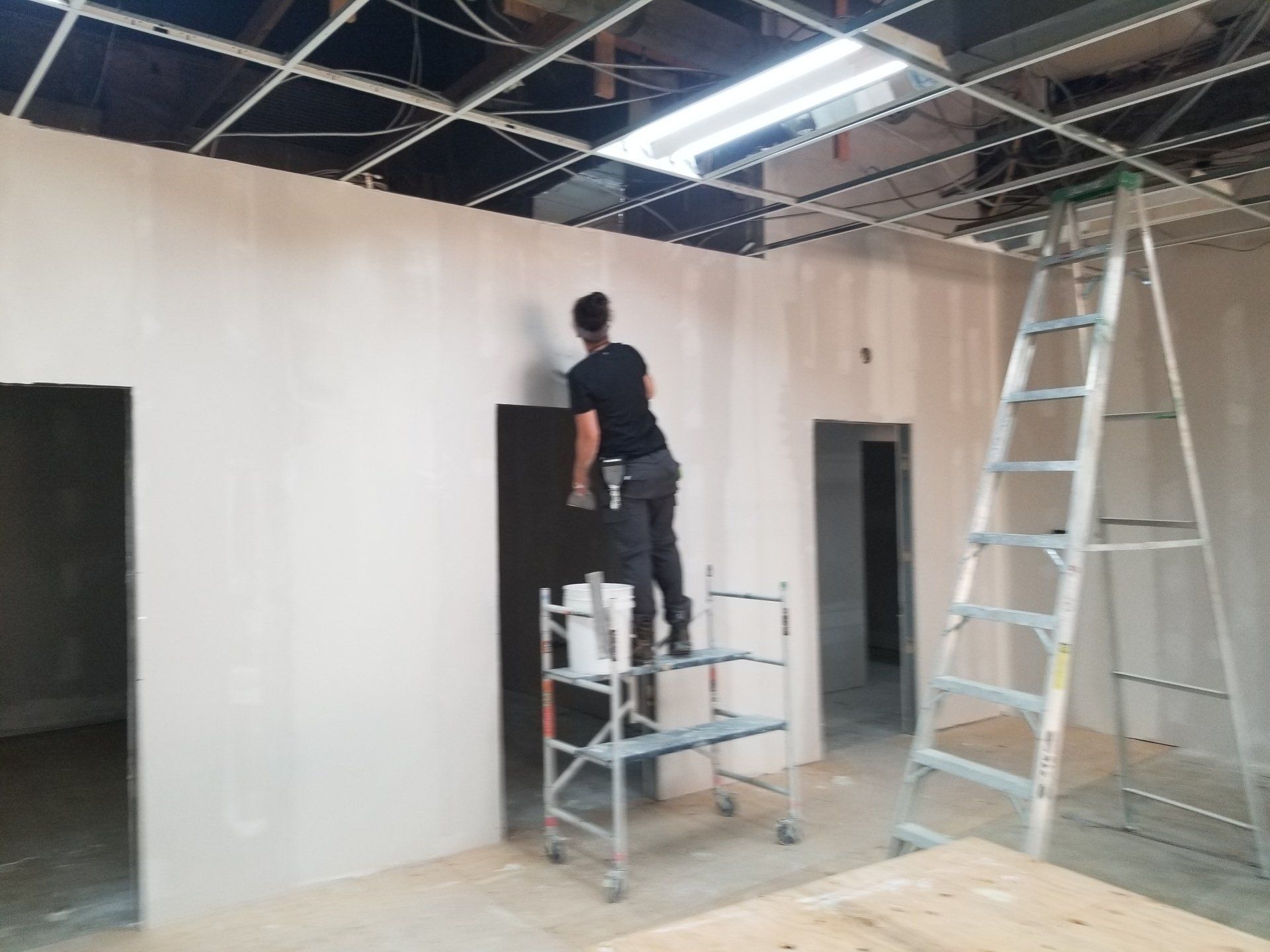 Un ouvrier peint un mur à l'intérieur d'une pièce, debout sur un échafaudage près d'une porte et d'une échelle.