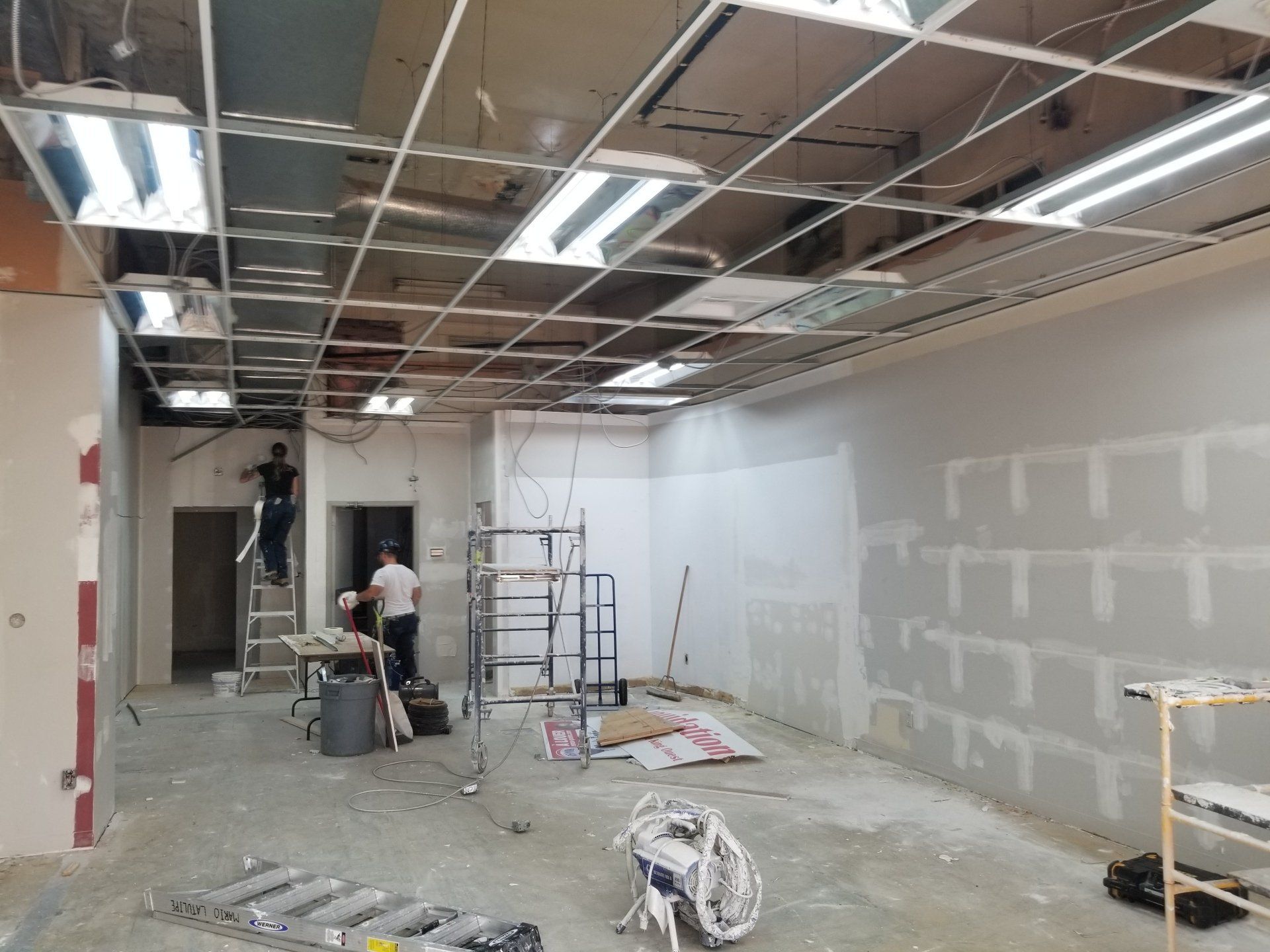 Chantier de rénovation avec deux ouvriers, échelles, outils et cloisons sèches non finies sous des luminaires apparents au plafond
