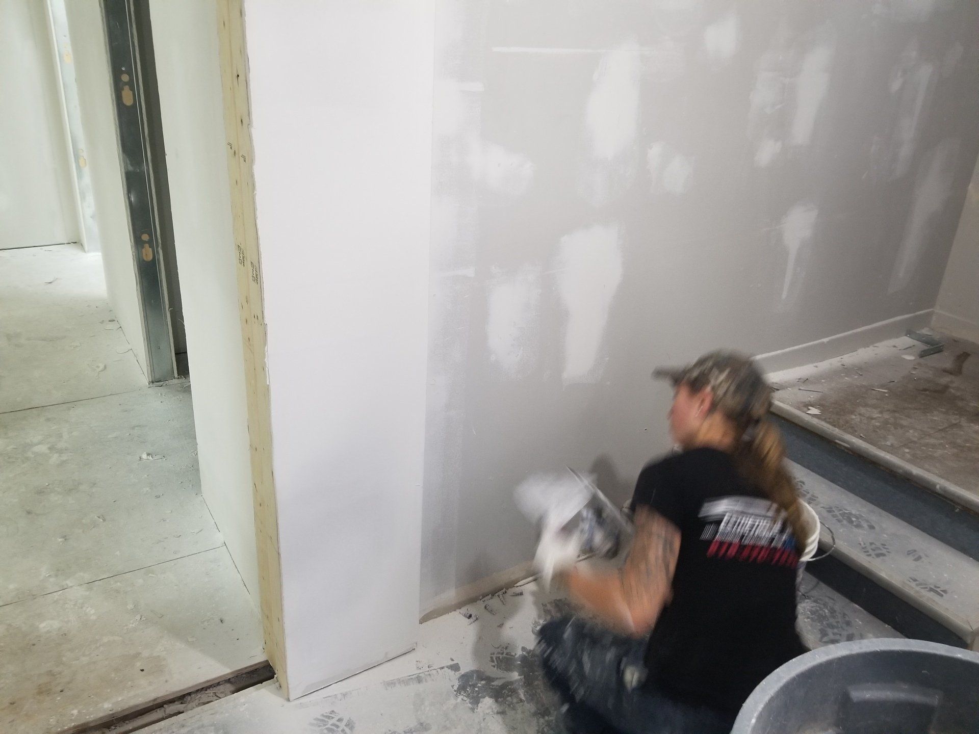 Une personne ponce des cloisons sèches dans une pièce en rénovation, avec des murs non finis et une porte à proximité.