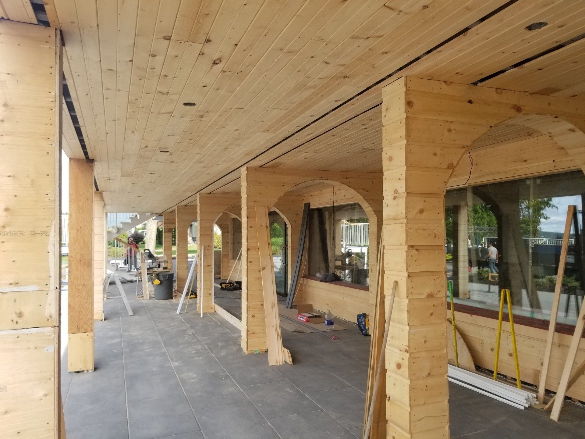 Passerelle couverte à ossature bois en construction, avec colonnes en bois, plafond en contreplaqué et ouvriers au loin.