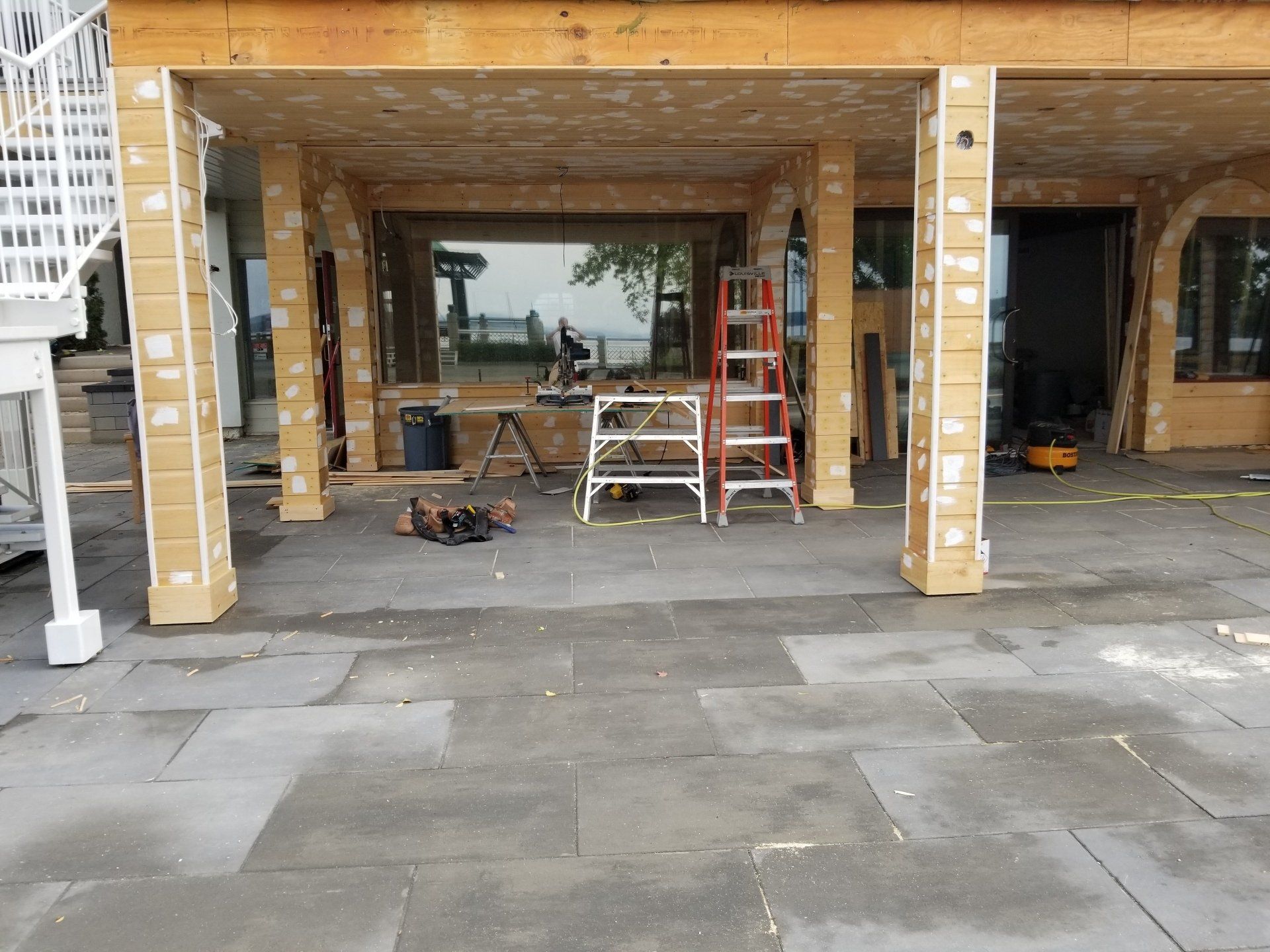 Terrasse extérieure en construction avec colonnes de briques, échelles, outils et pavés.