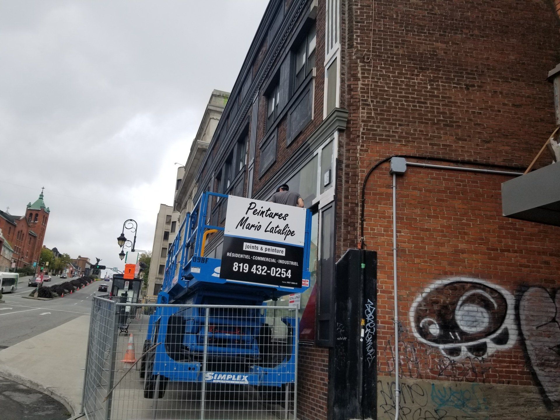 Ascenseur bleu stationné près de bâtiments en briques sur un trottoir de la ville, avec une enseigne commerciale.