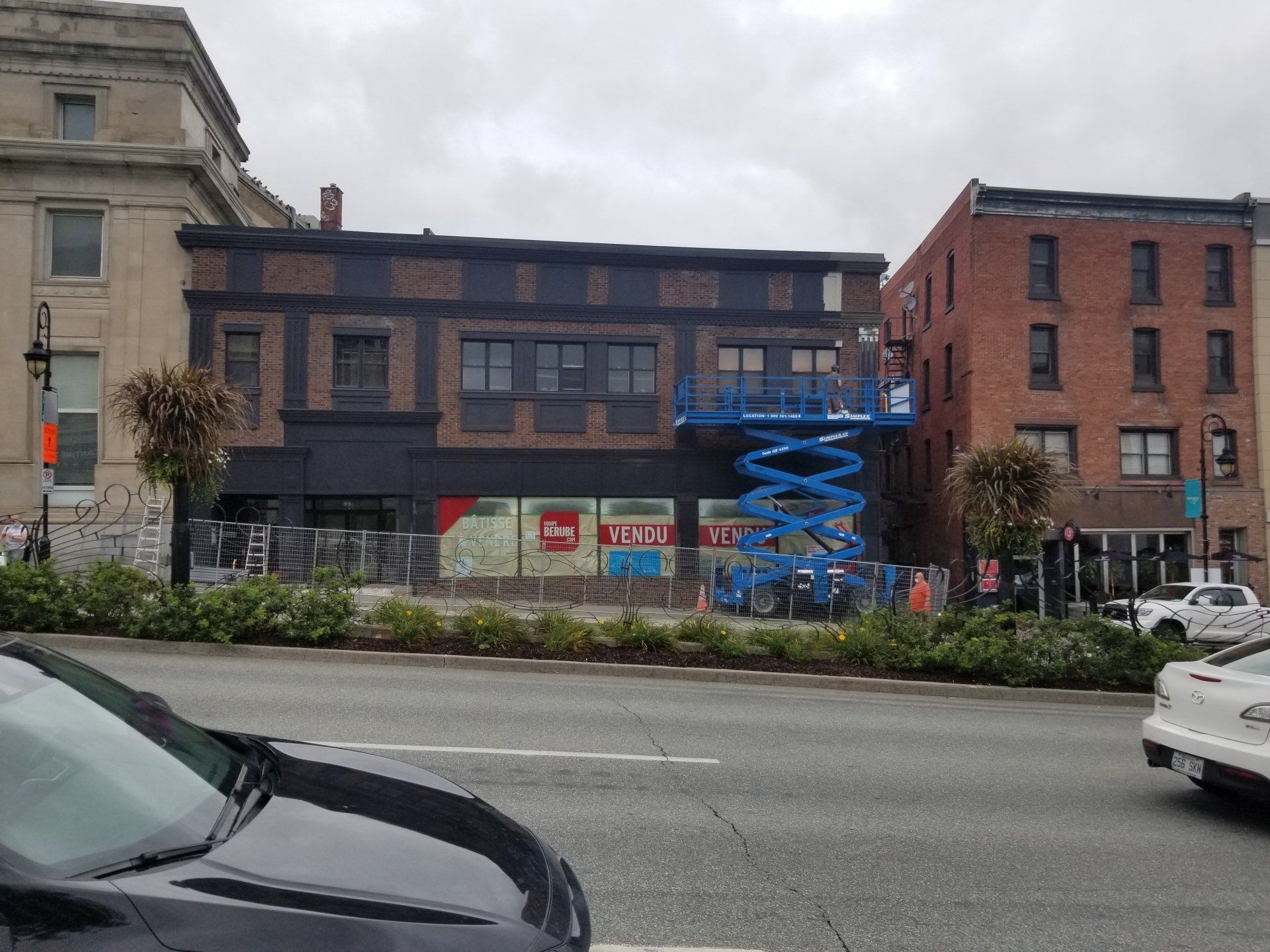 Rue urbaine avec une nacelle élévatrice bleue devant un immeuble en briques et une voiture blanche qui passe.