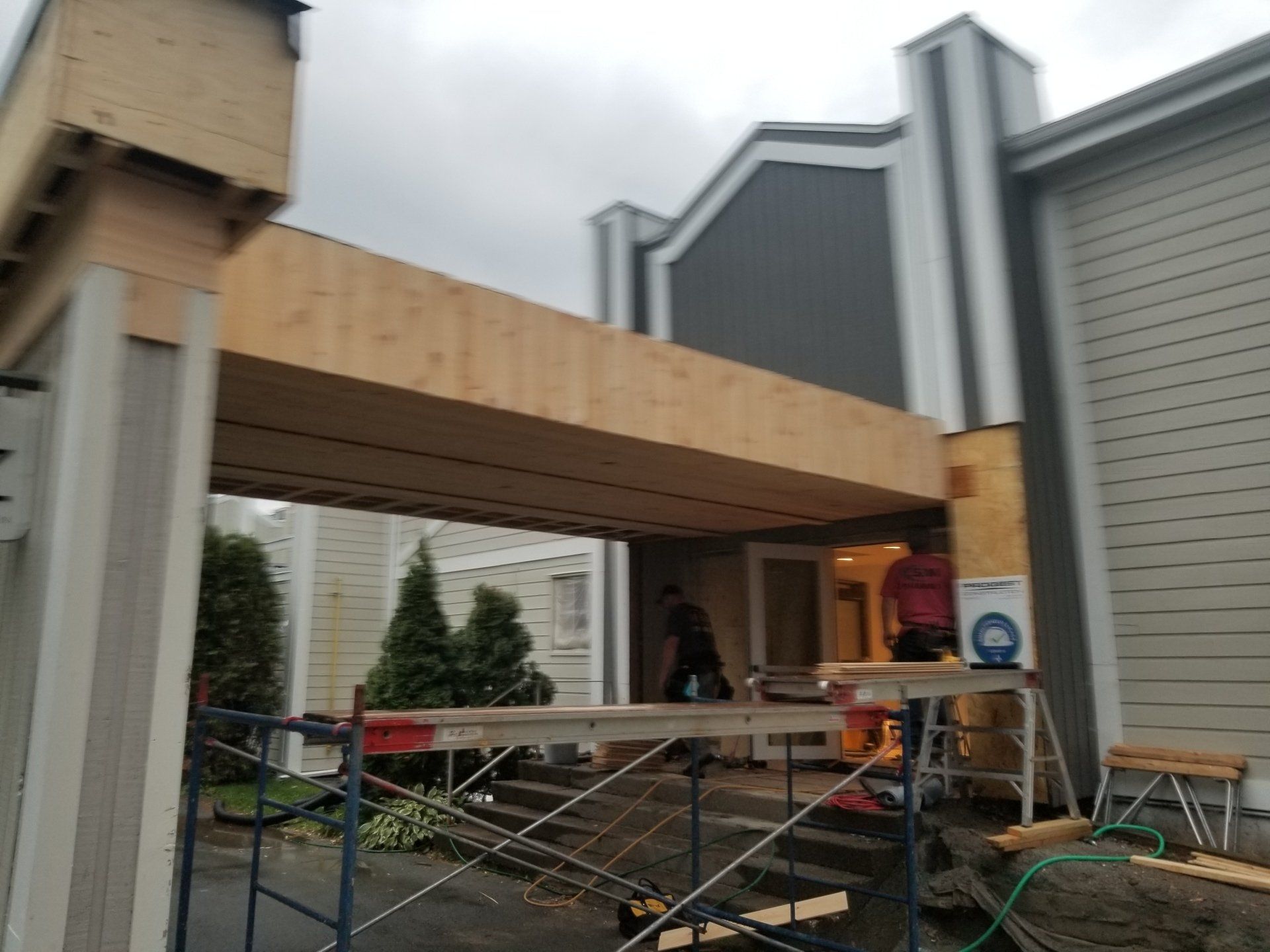 Entrée d'un bâtiment en construction avec un auvent en bois, des échafaudages et un ouvrier en arrière-plan.