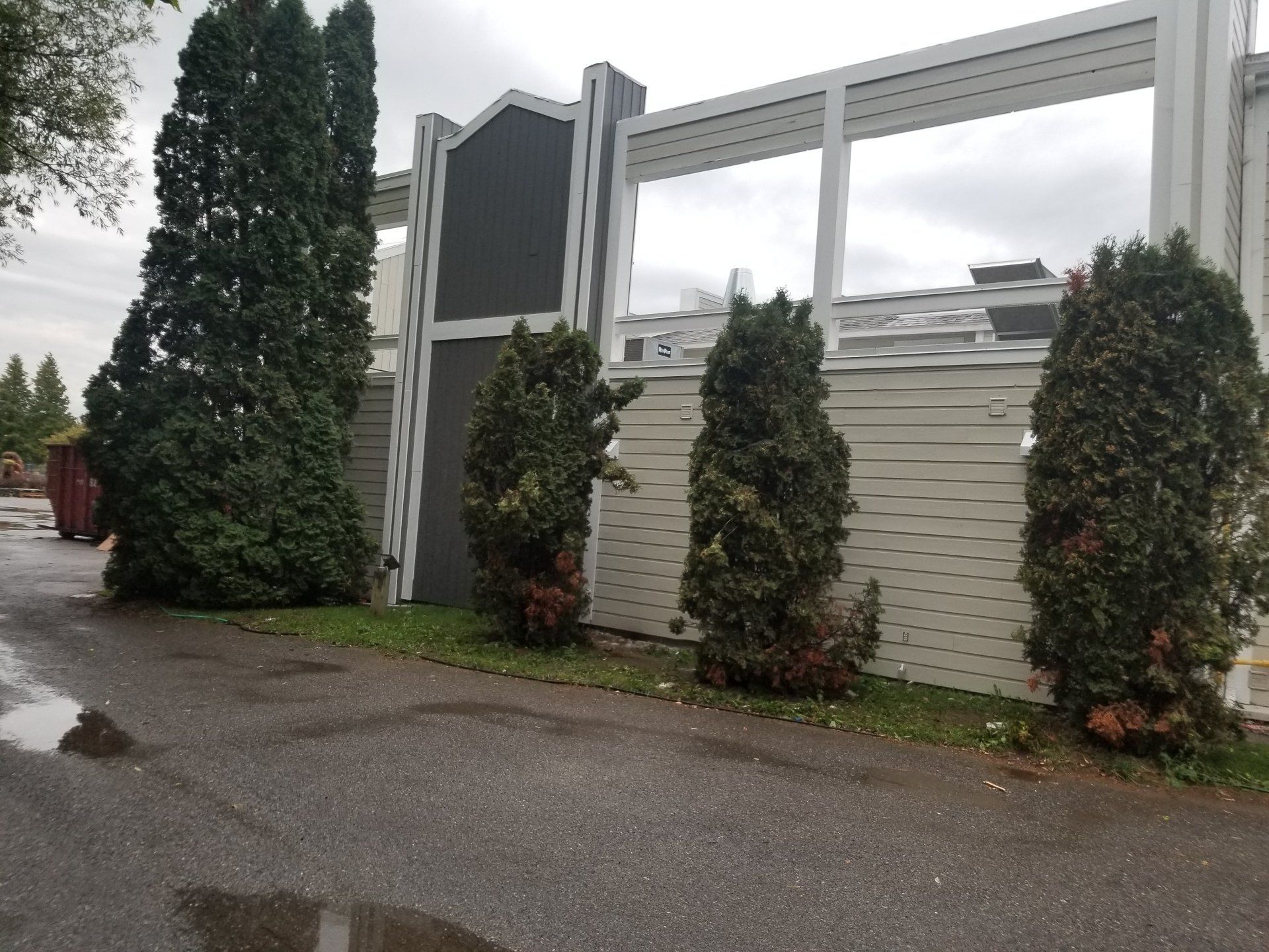 Portail d'entrée moderne avec panneaux blancs, grands arbres et allée mouillée sous un ciel nuageux