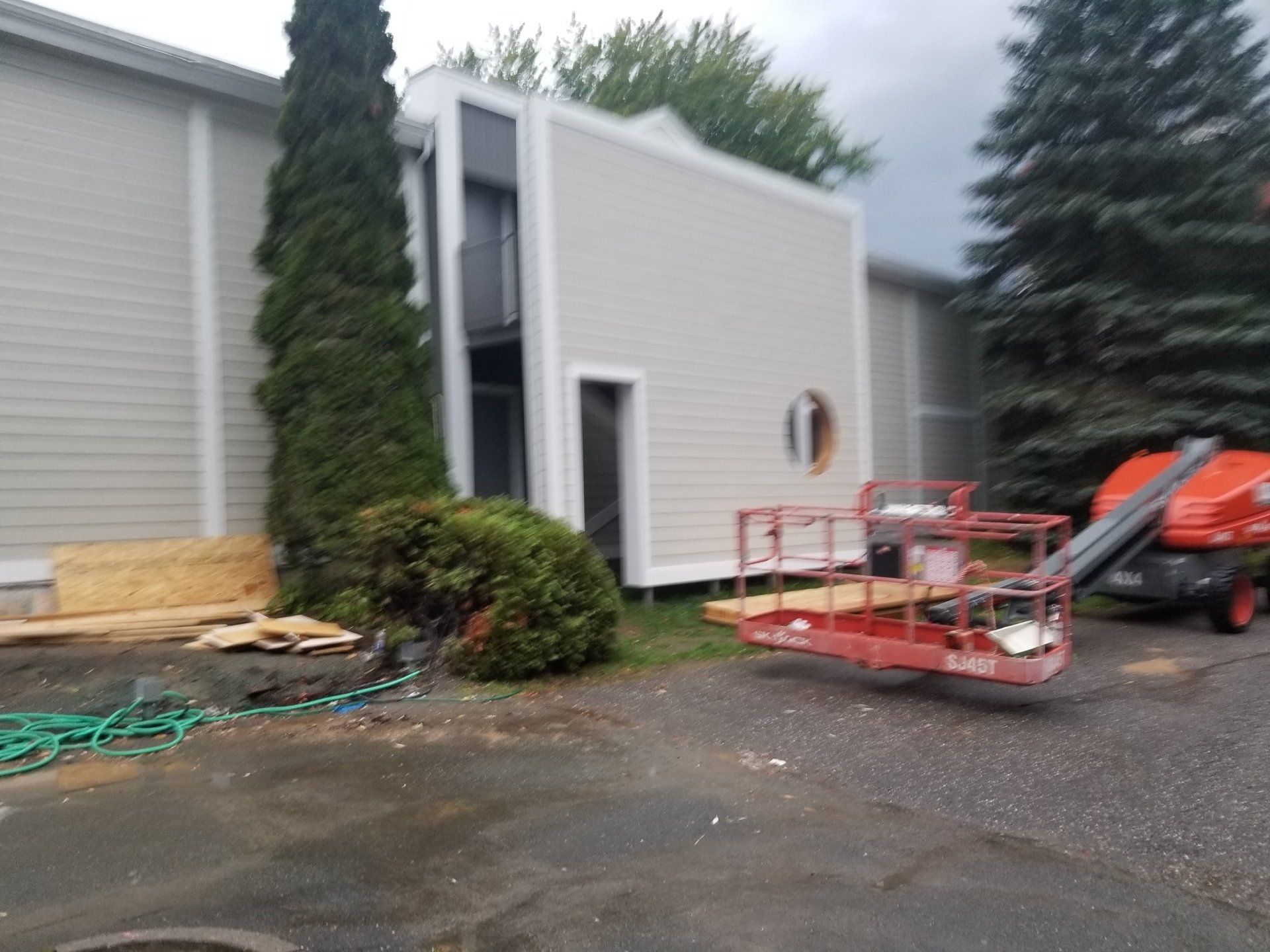 Extérieur d'un bâtiment avec de grands conifères, un ascenseur rouge et des matériaux de construction sur un terrain pavé humide
