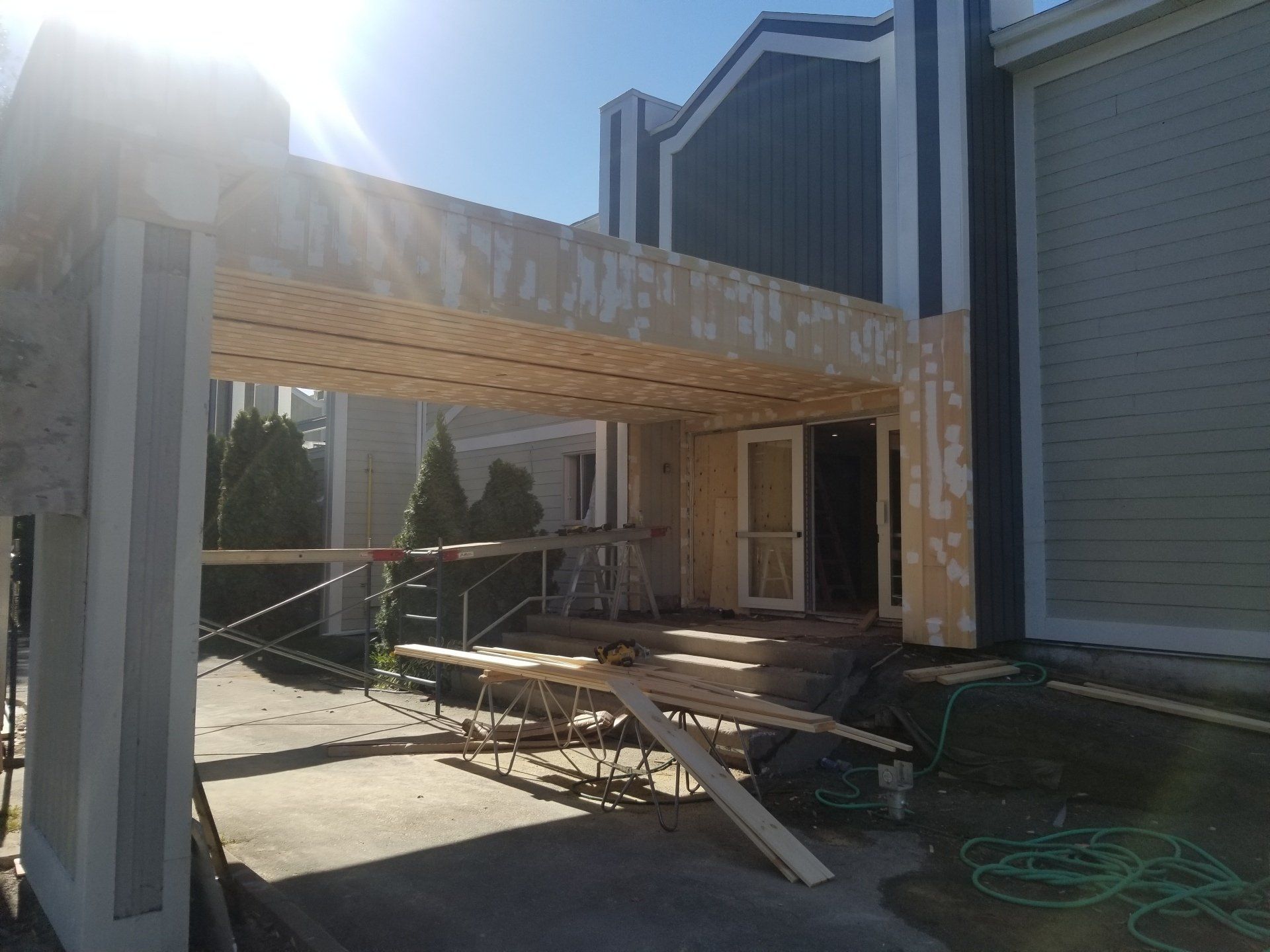 Pergola en bois partiellement construite, adossée à une maison, avec des matériaux de construction et une échelle dans la terrasse.