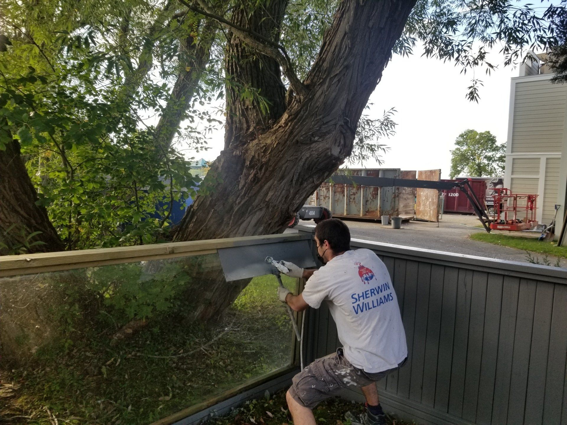 Une personne est accroupie sur une terrasse, ajustant une rambarde en verre à côté d'un grand arbre et d'une maison.
