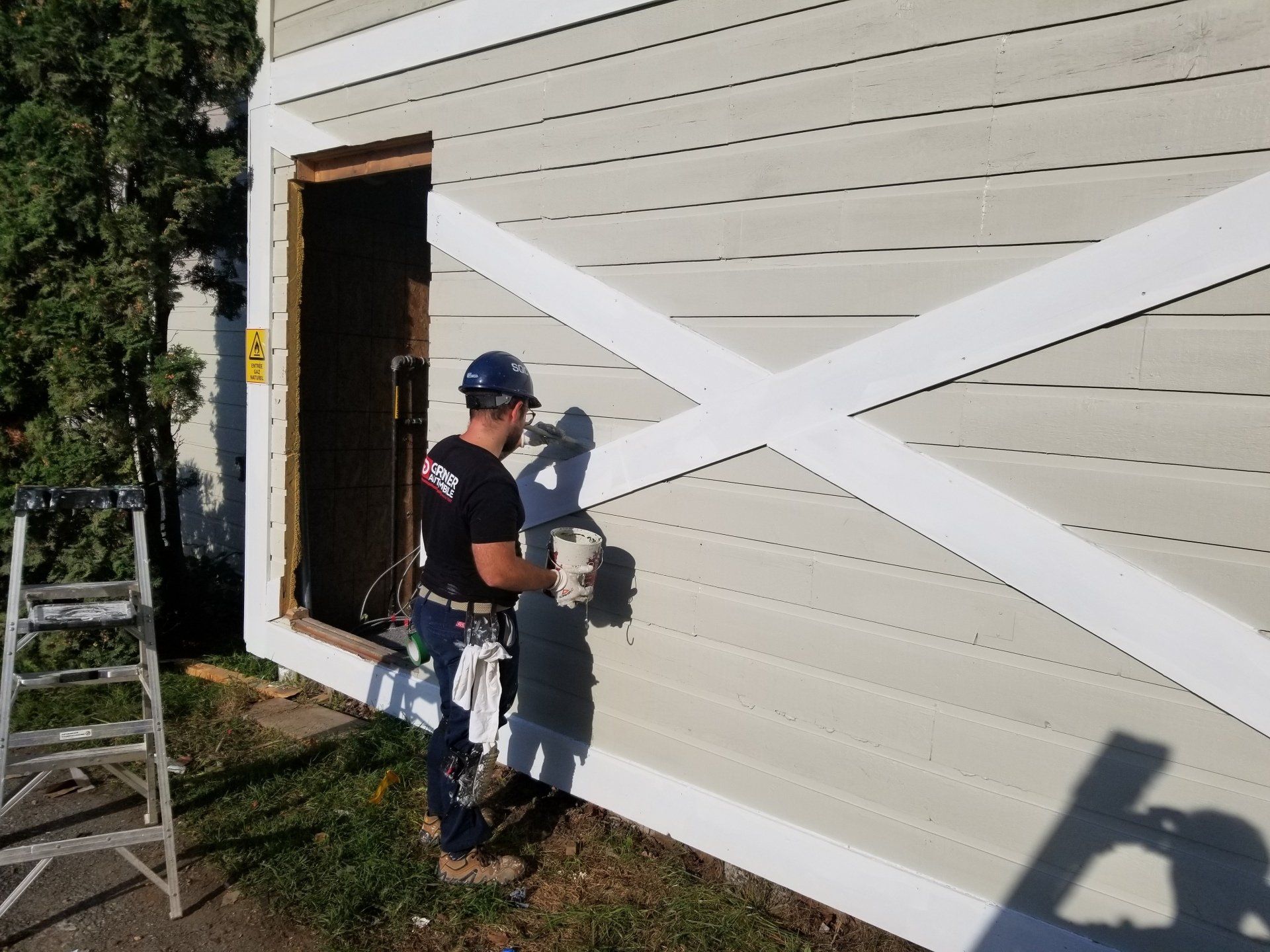 Un ouvrier peint un mur extérieur avec des entretoises blanches à côté d'une fenêtre ouverte et d'une échelle