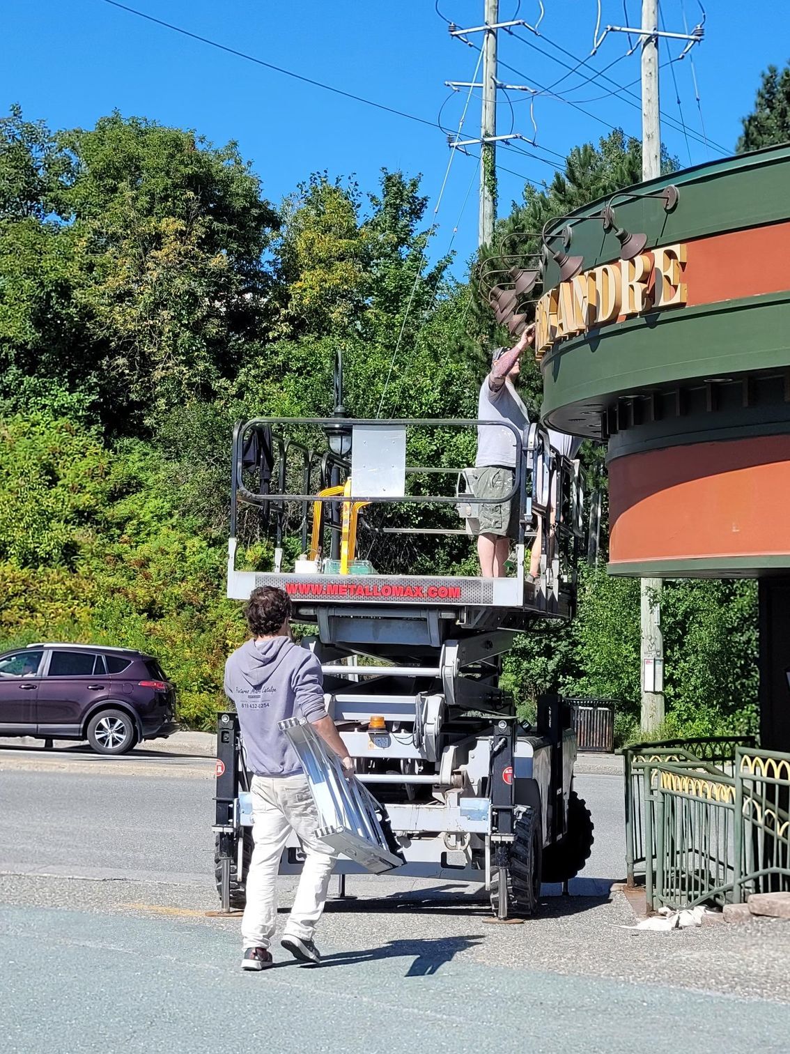 Une personne charge du matériel sur un camion à côté d'un bâtiment vert, au bord d'une route ensoleillée.