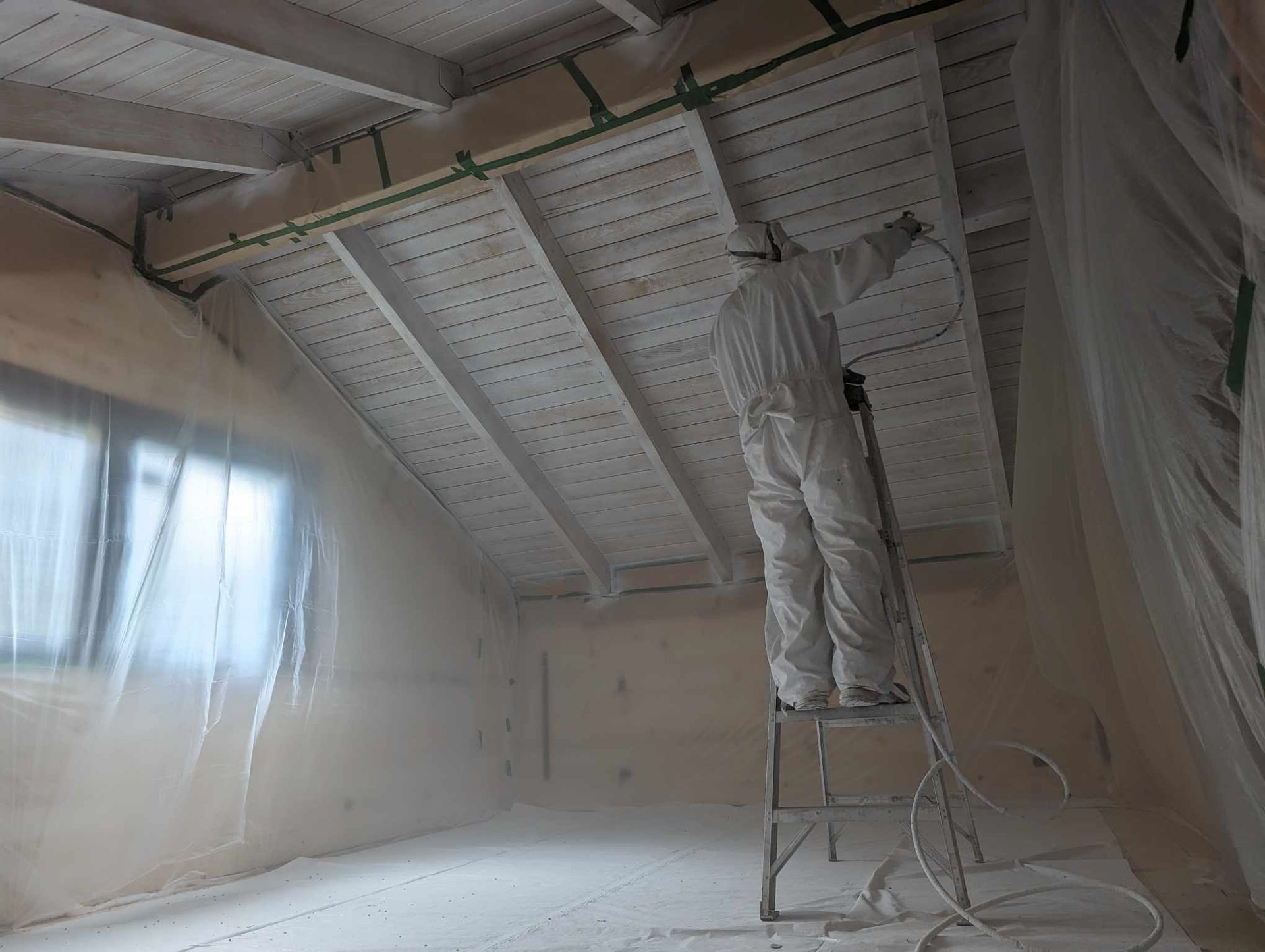 Un ouvrier peint un plafond du haut d'une échelle dans une pièce recouverte d'une bâche de protection en plastique.