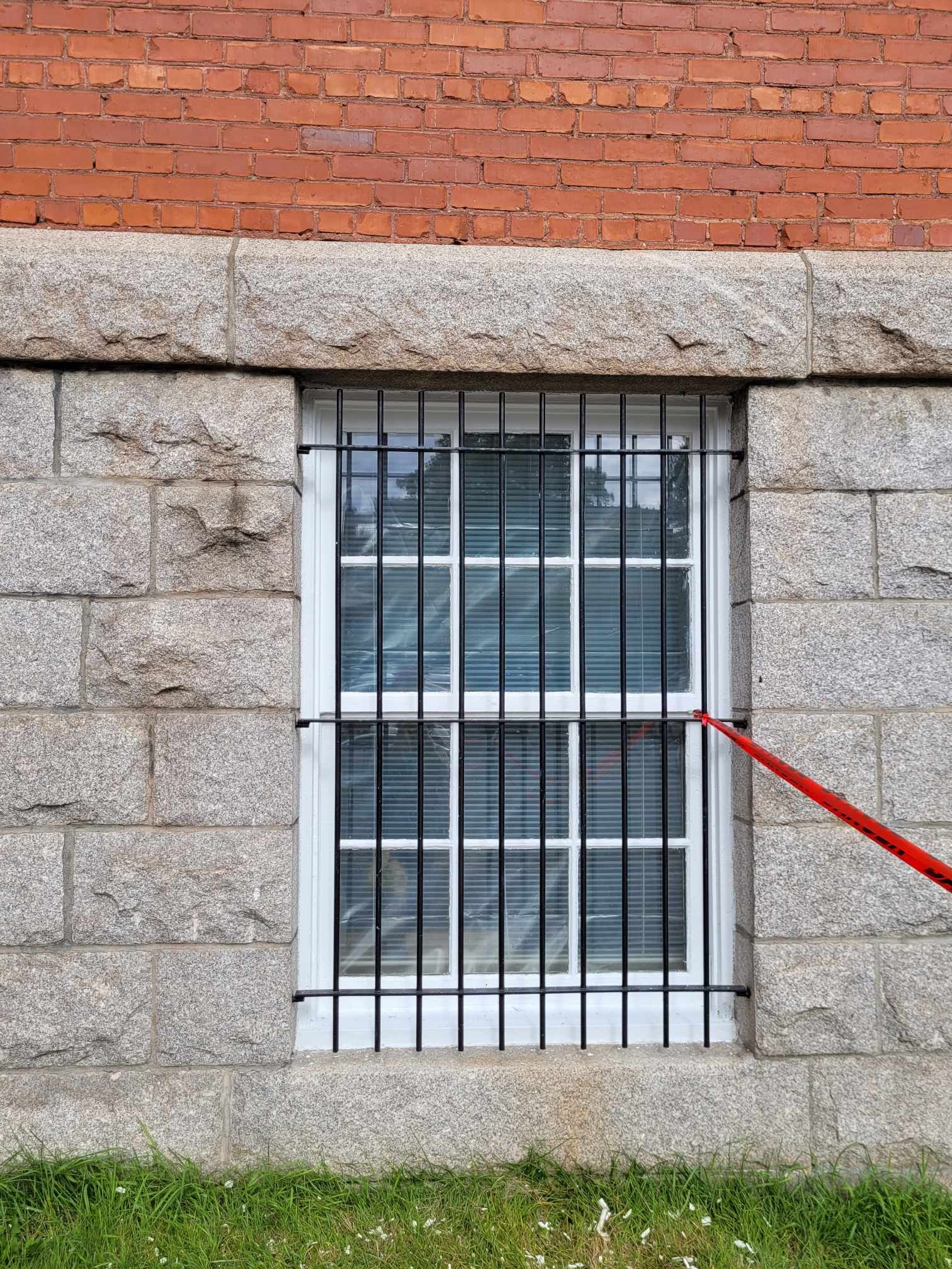 Fenêtre de cave à barreaux dans un mur de pierre grise sous des briques rouges, avec une flèche rouge pointant vers le bas à droite