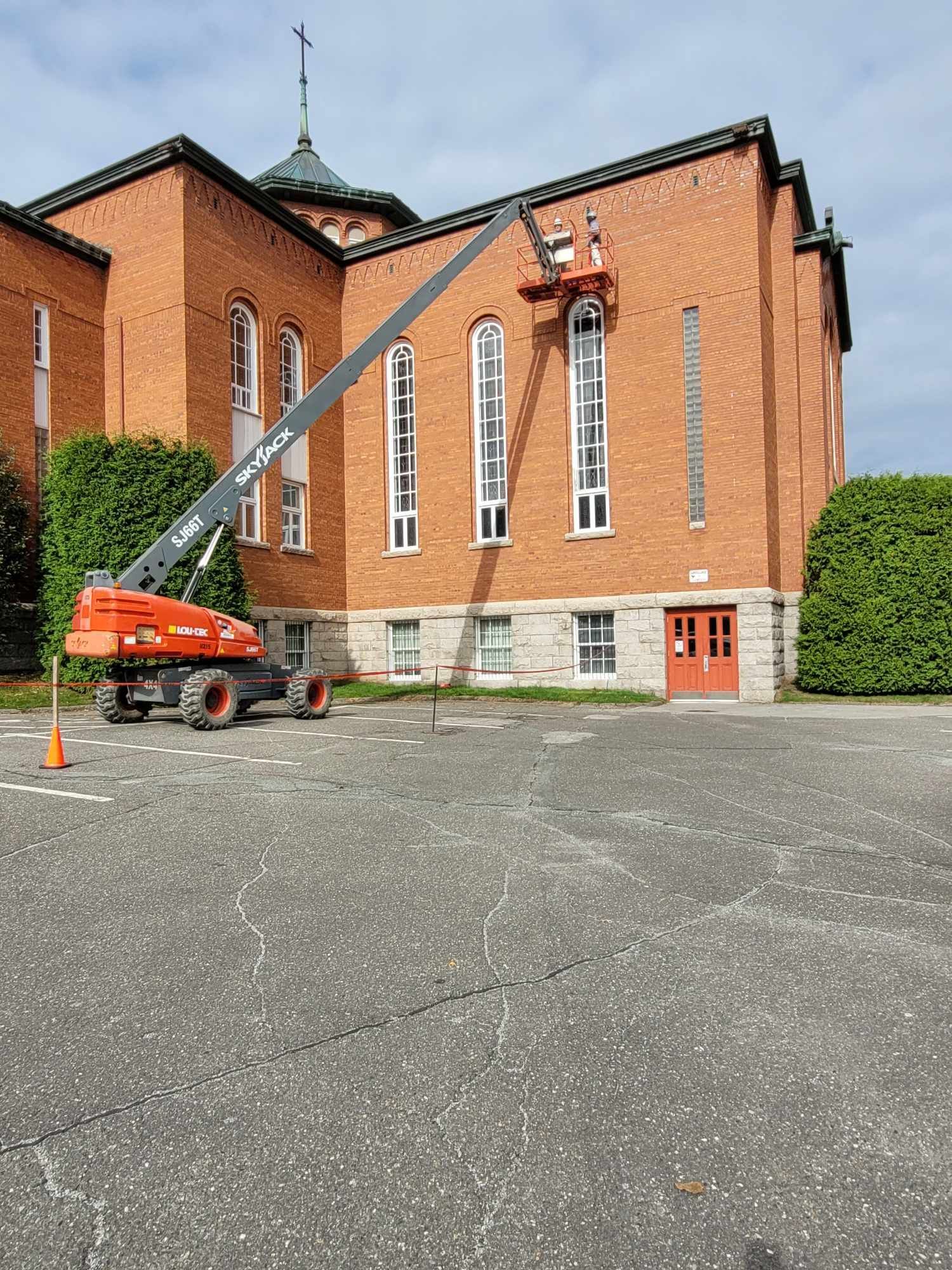 Une nacelle élévatrice soulève des oranges à côté d'un bâtiment en briques rouges au toit en pente et au mur latéral bordé de haies.