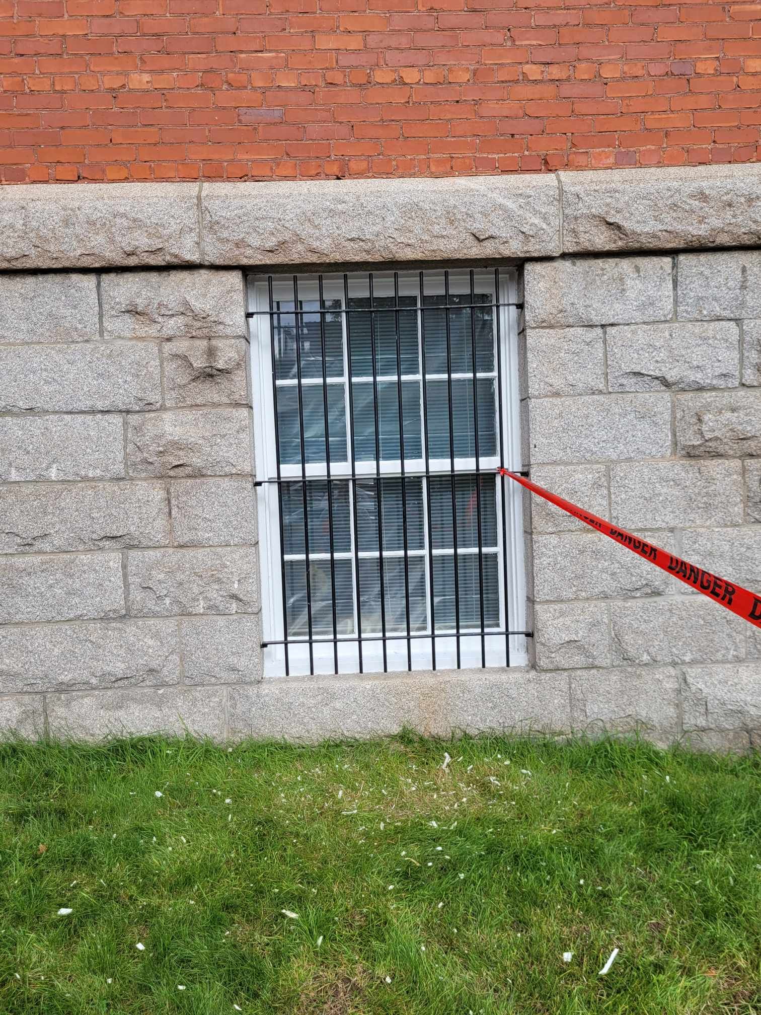 Fenêtre de cave grillagée dans un mur de pierre, avec une laisse ou une sangle rouge pointée vers elle.