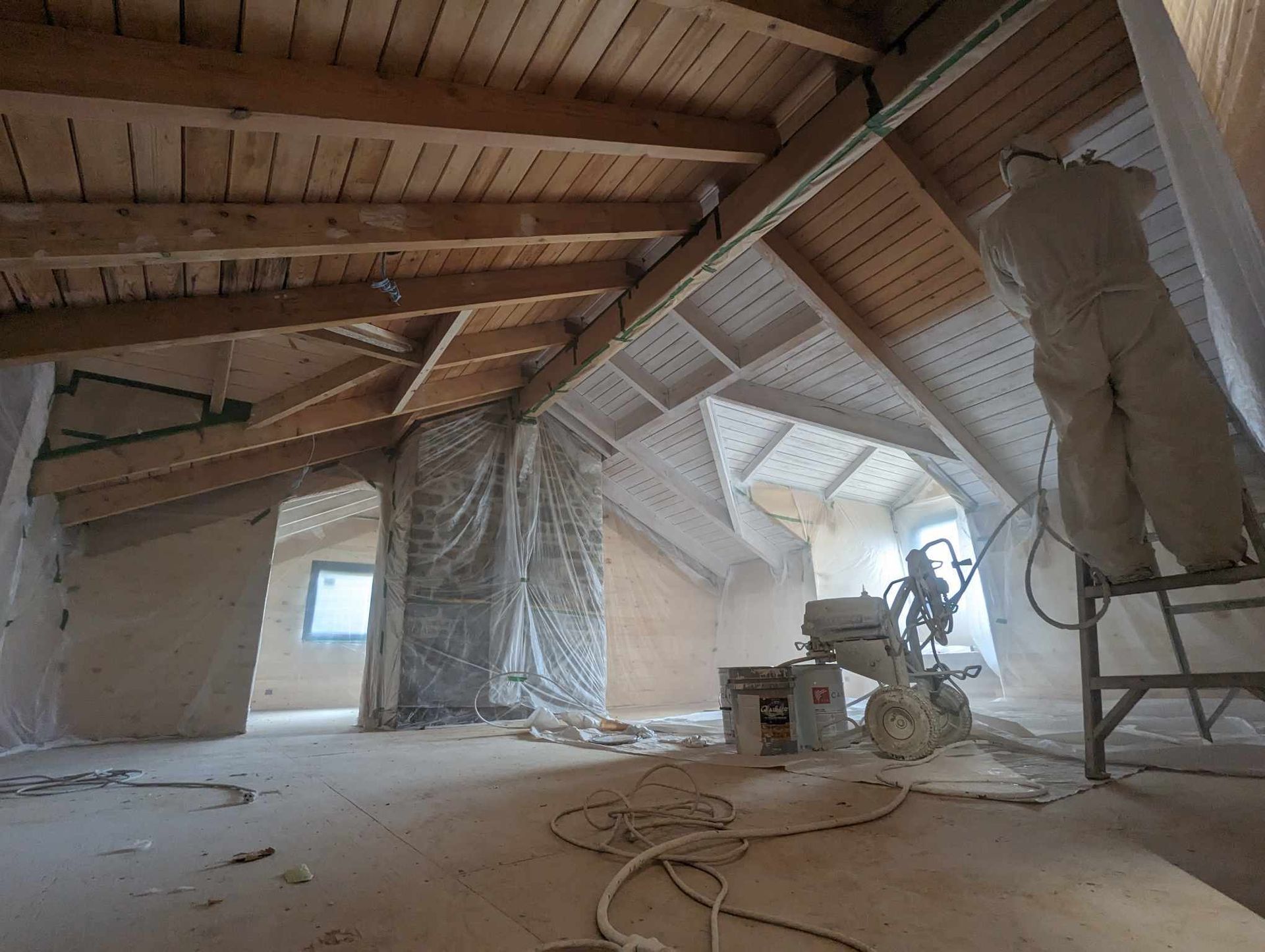 Grenier poussiéreux en cours de rénovation, avec poutres en bois apparentes, bâches en plastique suspendues et outils de construction.