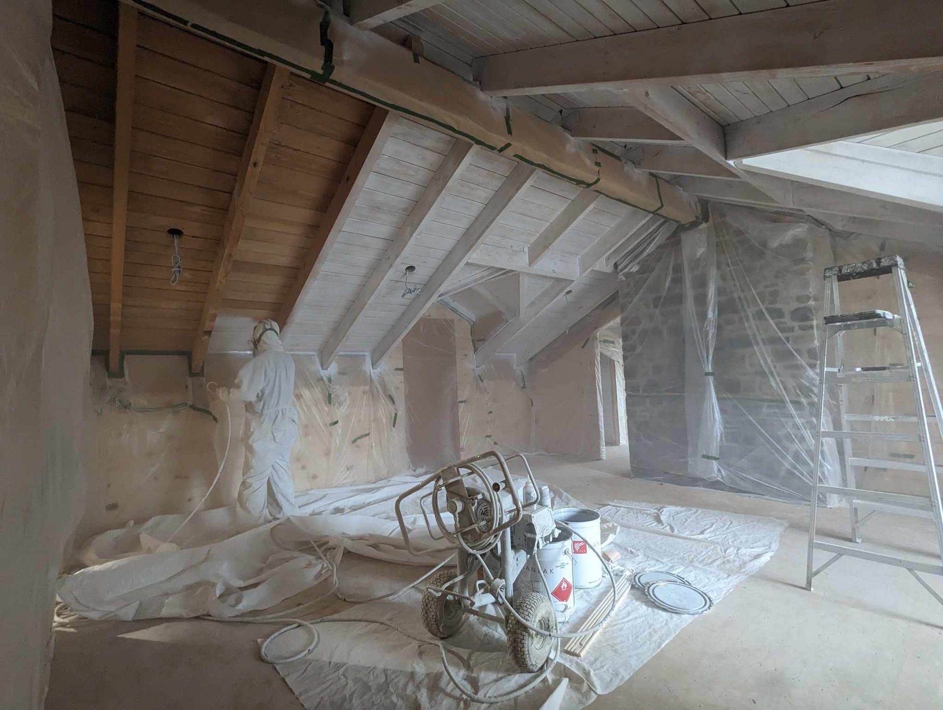 Grenier poussiéreux en cours de rénovation, avec poutres apparentes, isolation, outils et une échelle appuyée contre le mur.