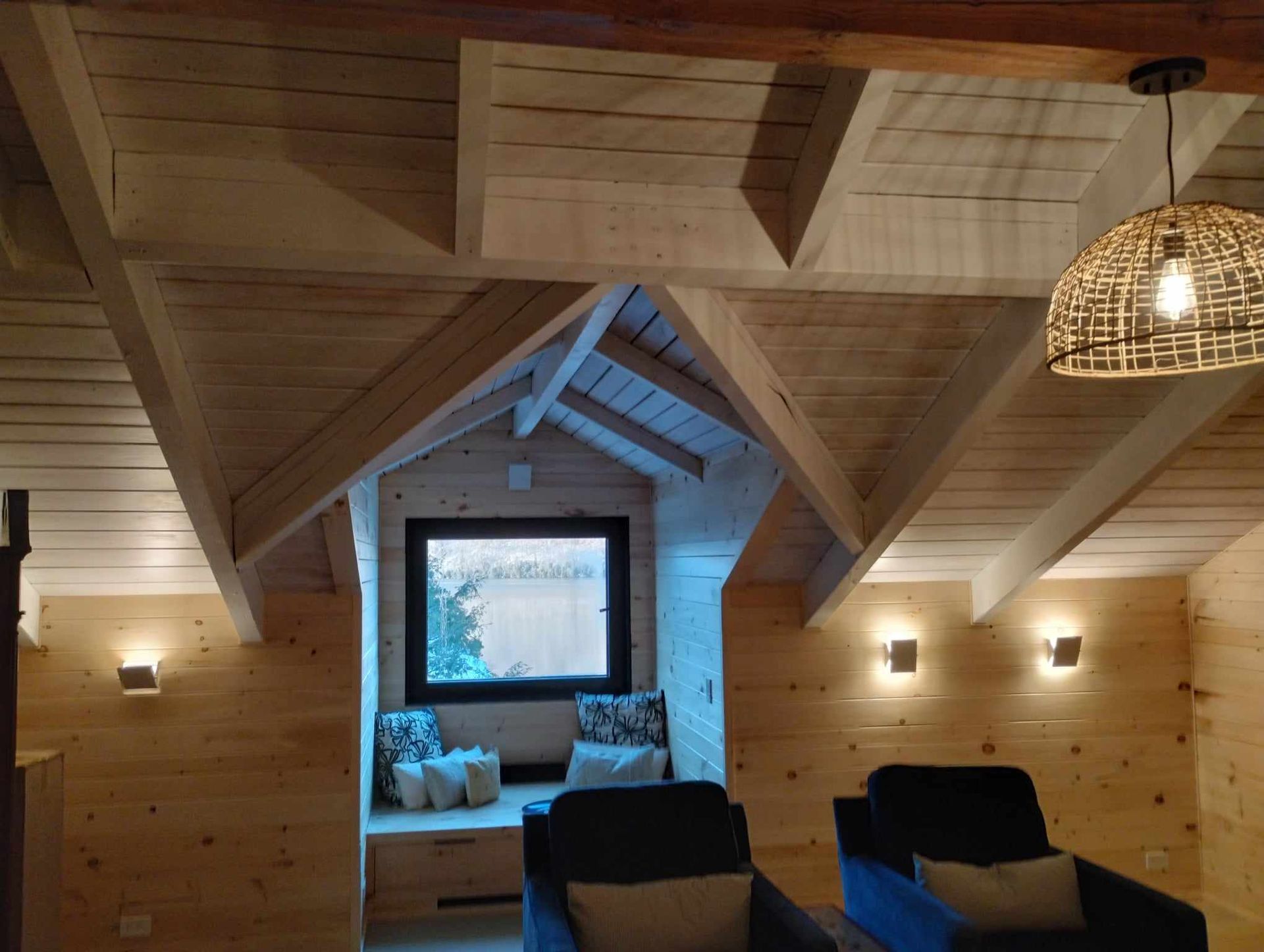 Chambre mansardée confortable en bois avec banquette près de la fenêtre, deux chaises et une lampe suspendue en osier.