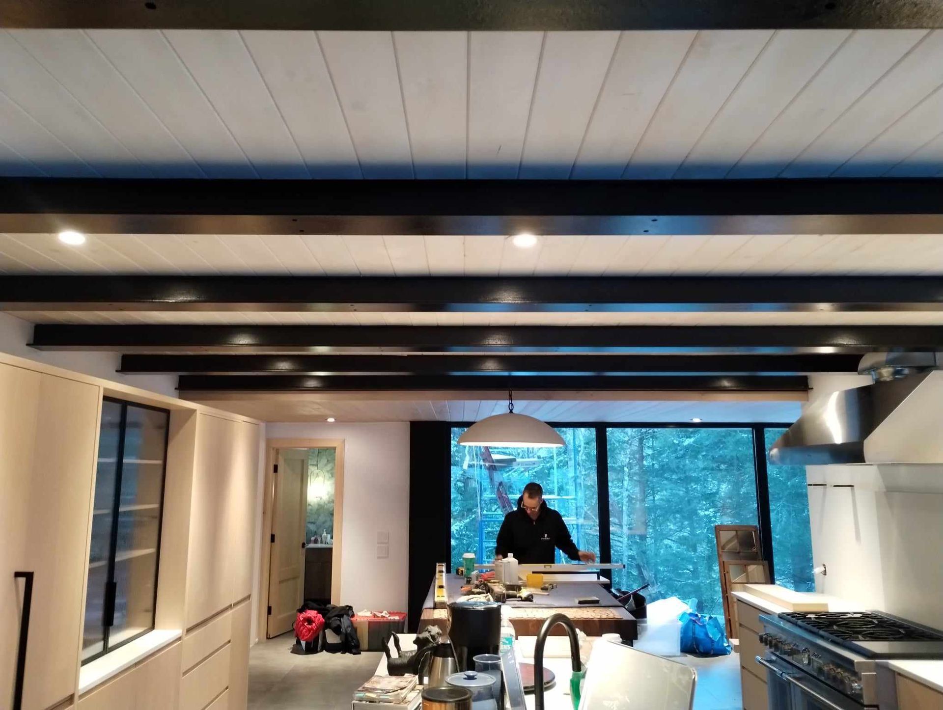 Intérieur de cuisine moderne avec poutres apparentes et une personne travaillant à l'îlot près d'une grande fenêtre