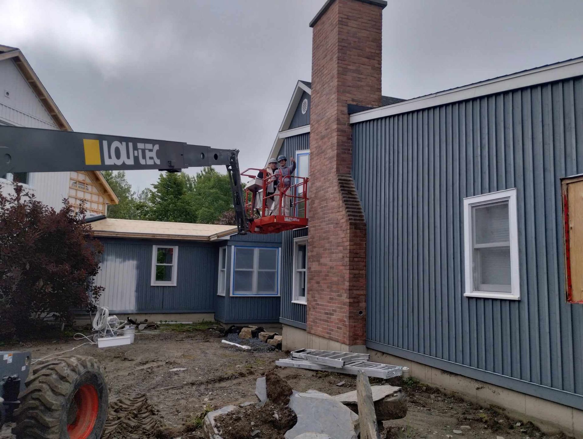 Un monte-charge de chantier à côté d'une maison bleue avec une cheminée en briques et des ouvriers sur une plateforme surélevée