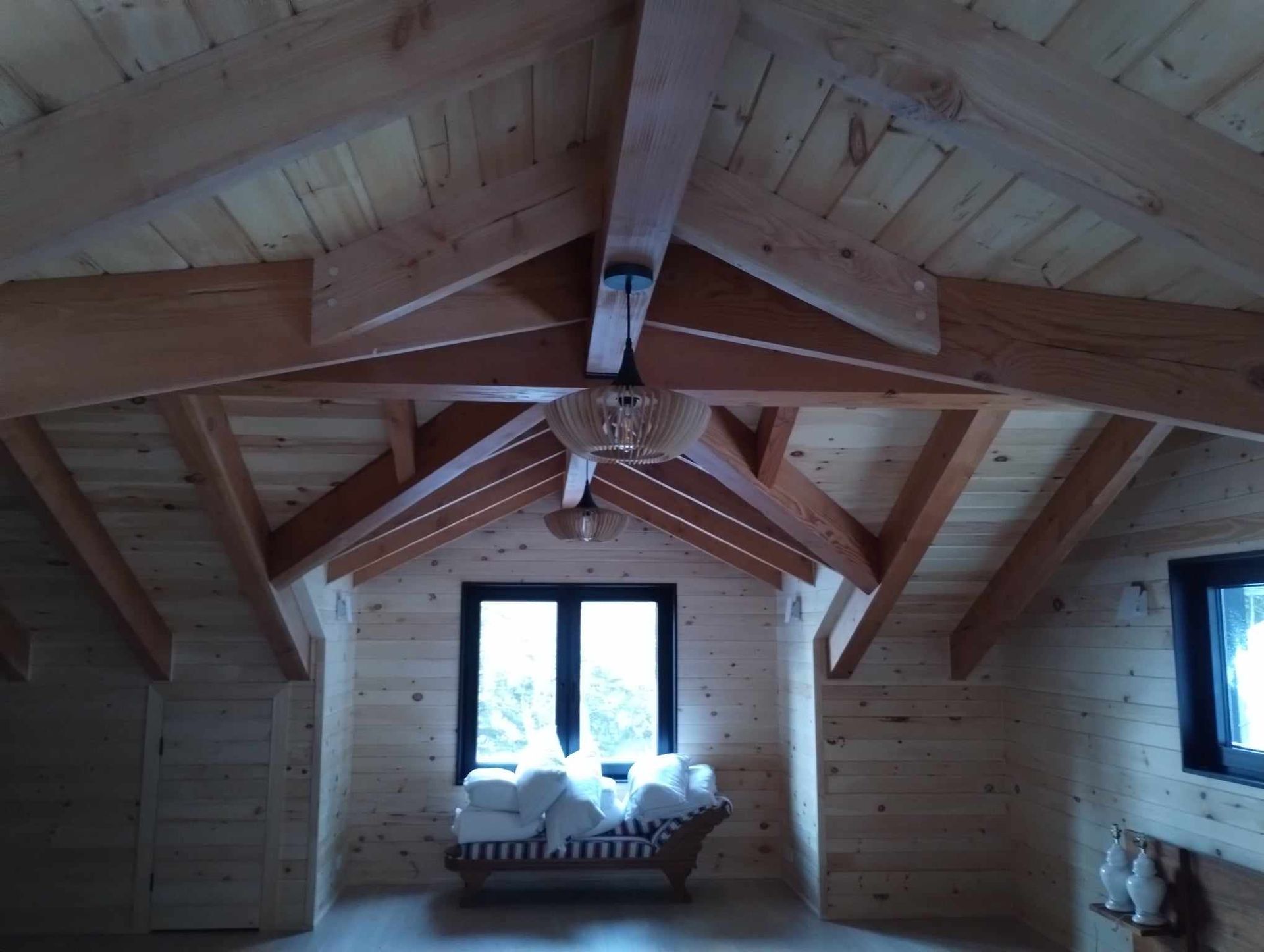 Pièce mansardée en bois avec poutres apparentes, banquette près de la fenêtre et douce lumière naturelle.
